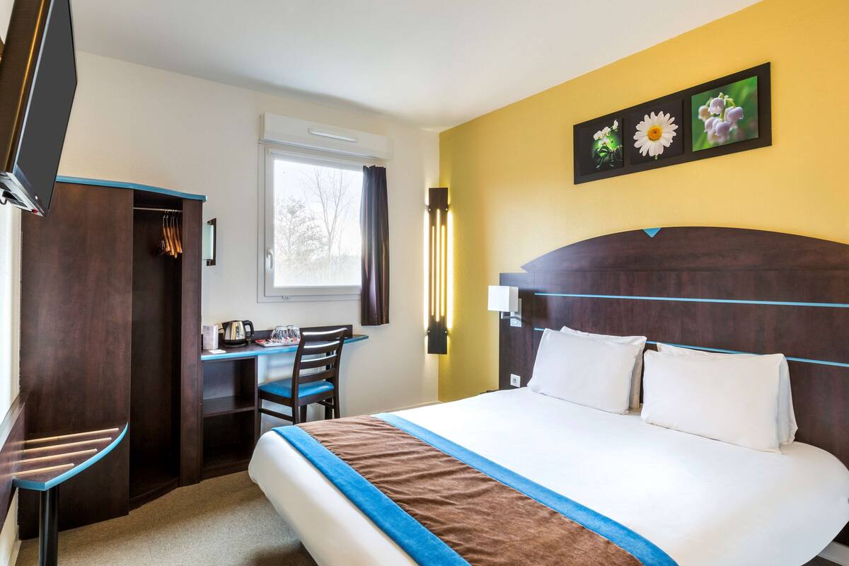 A hotel room with a bed, a desk, and a chair. There is a window with curtains, a wall-mounted picture, and a bedside table.