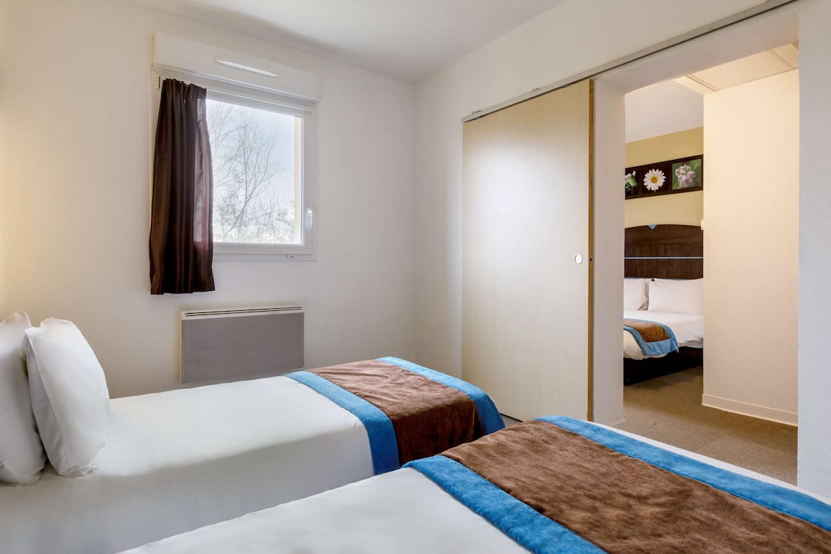 A hotel room with two beds, a window with curtains, a radiator, and a sliding door leading to another room.