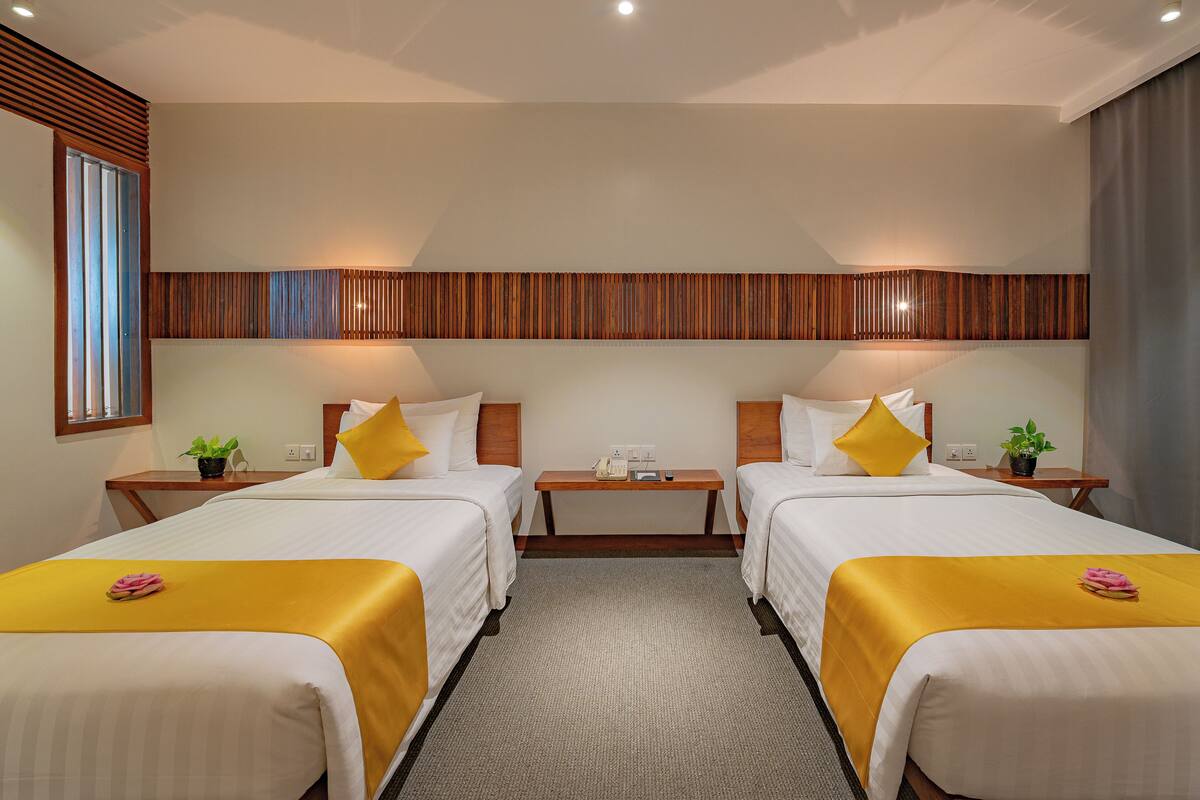 A hotel room with two beds, each with a yellow bedspread and white pillows, a wooden headboard, bedside tables with lamps, and a small potted plant on each table.
