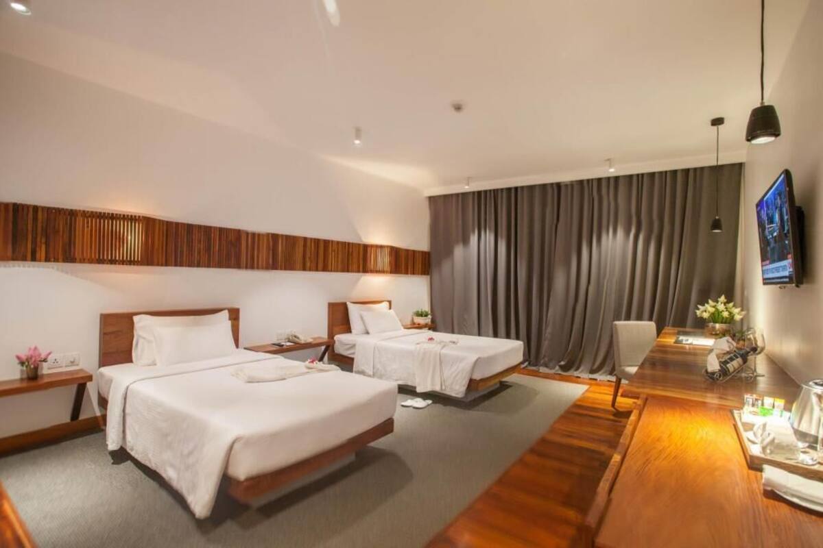 A hotel room with two beds, a wooden headboard, a flat-screen TV, a desk, and a dining table.