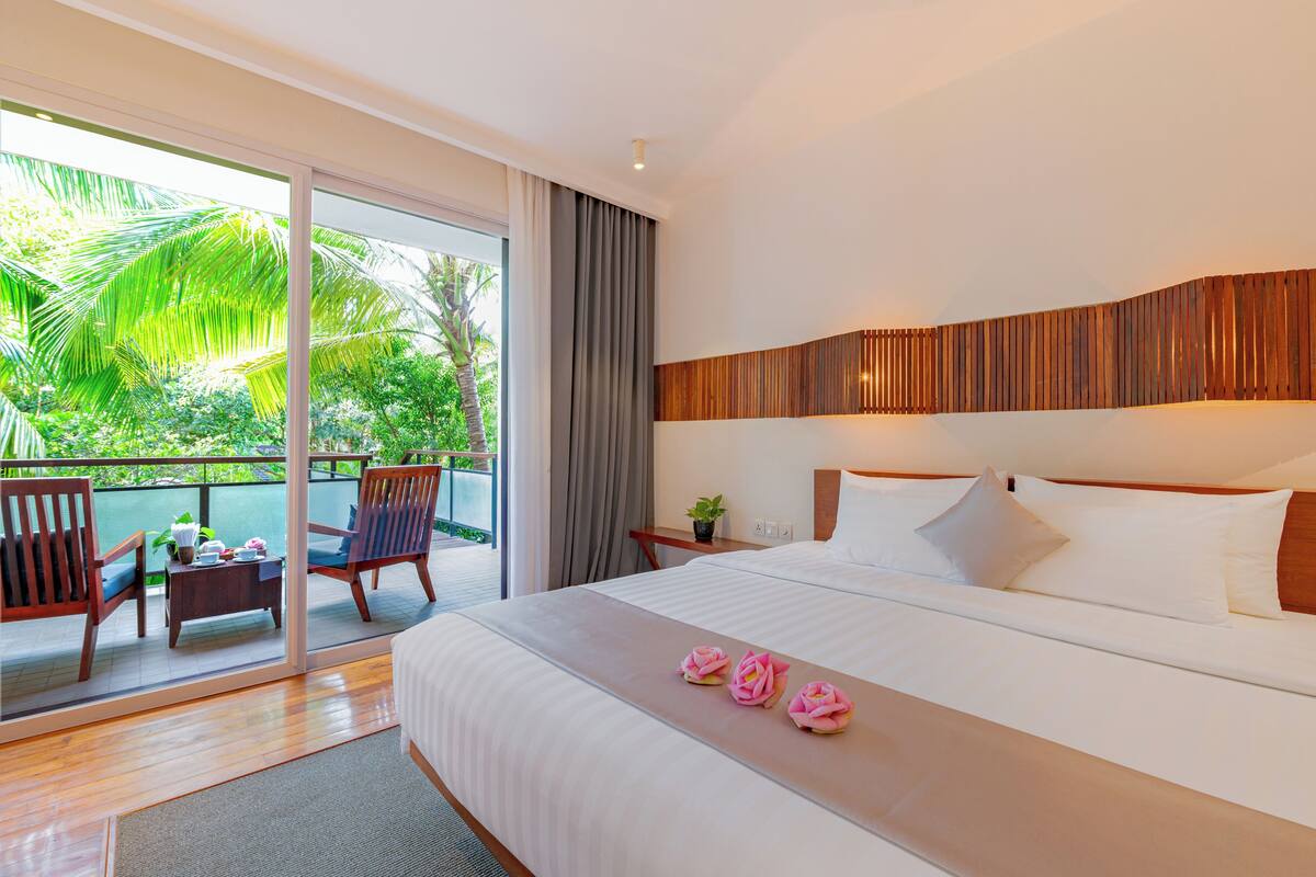 A hotel room with a large bed, a balcony with chairs, and a view of greenery.