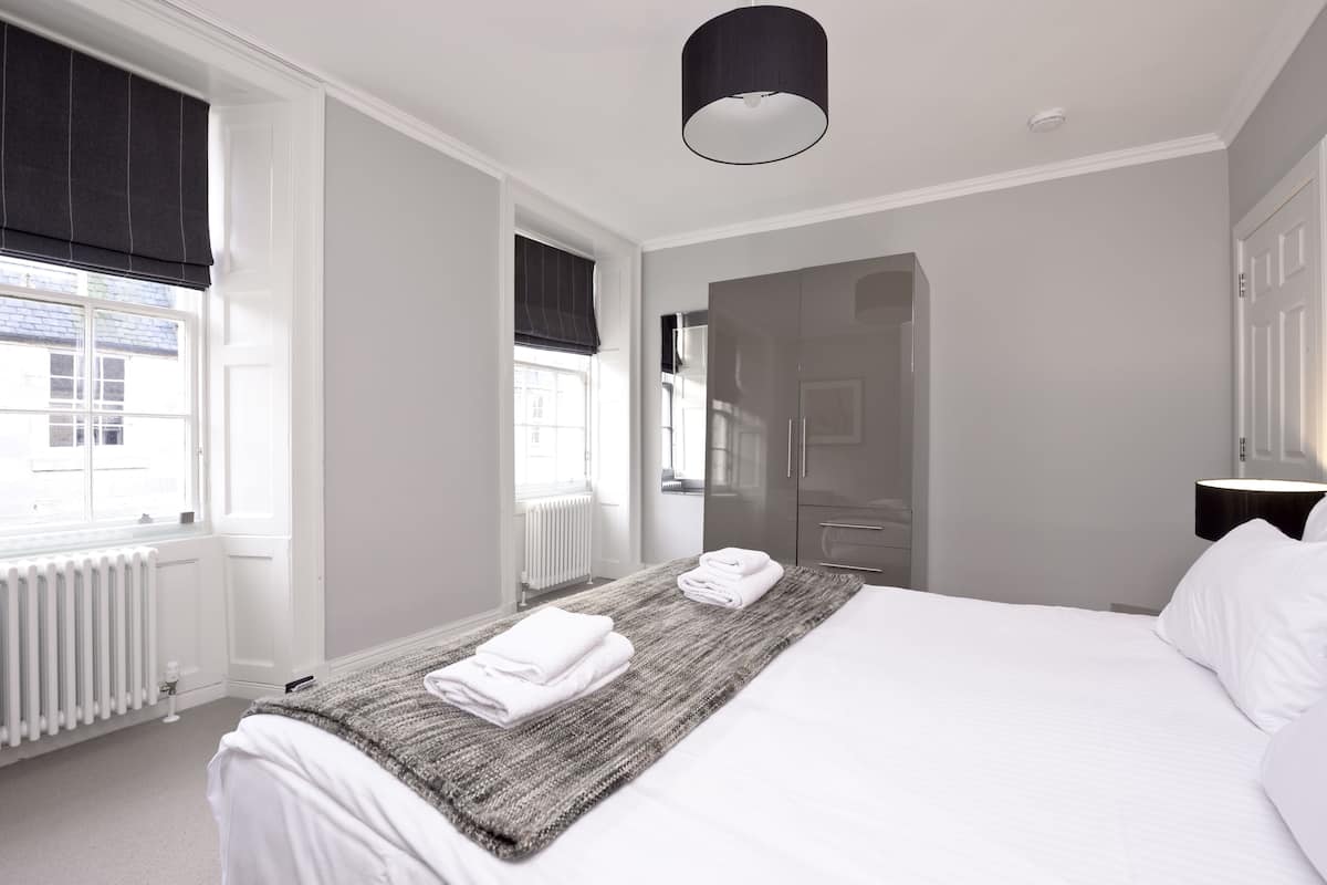Une chambre d’hôtel moderne, dotée d’un grand lit, d’un radiateur, d’une fenêtre avec stores et d’une porte donnant sur une autre pièce.