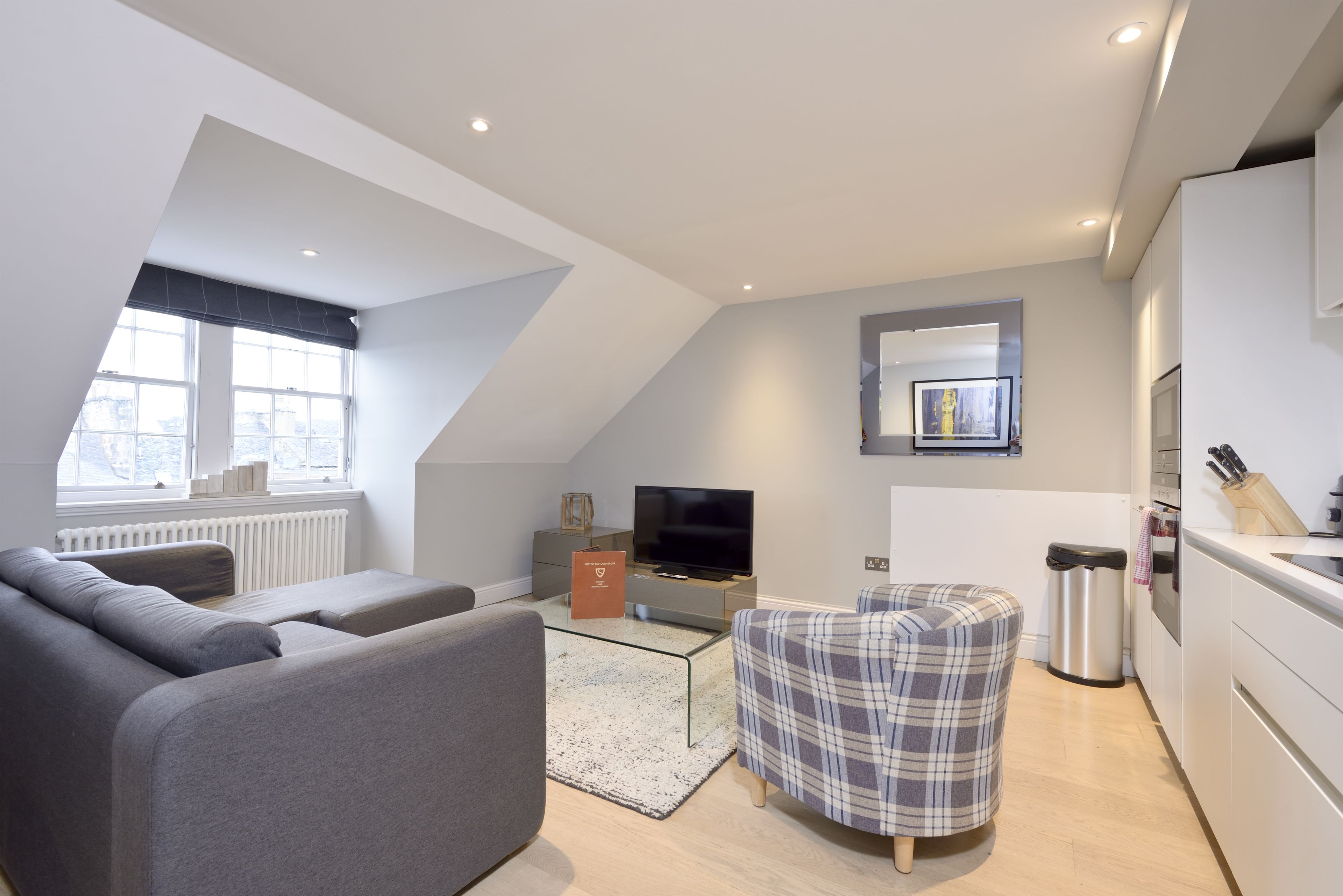 Un loft moderno con sala de estar que incluye un sofá gris, un sillón escocés, una mesa de centro de vidrio y un televisor de pantalla plana. La cocina tiene armarios blancos, una encimera y un ventanal.