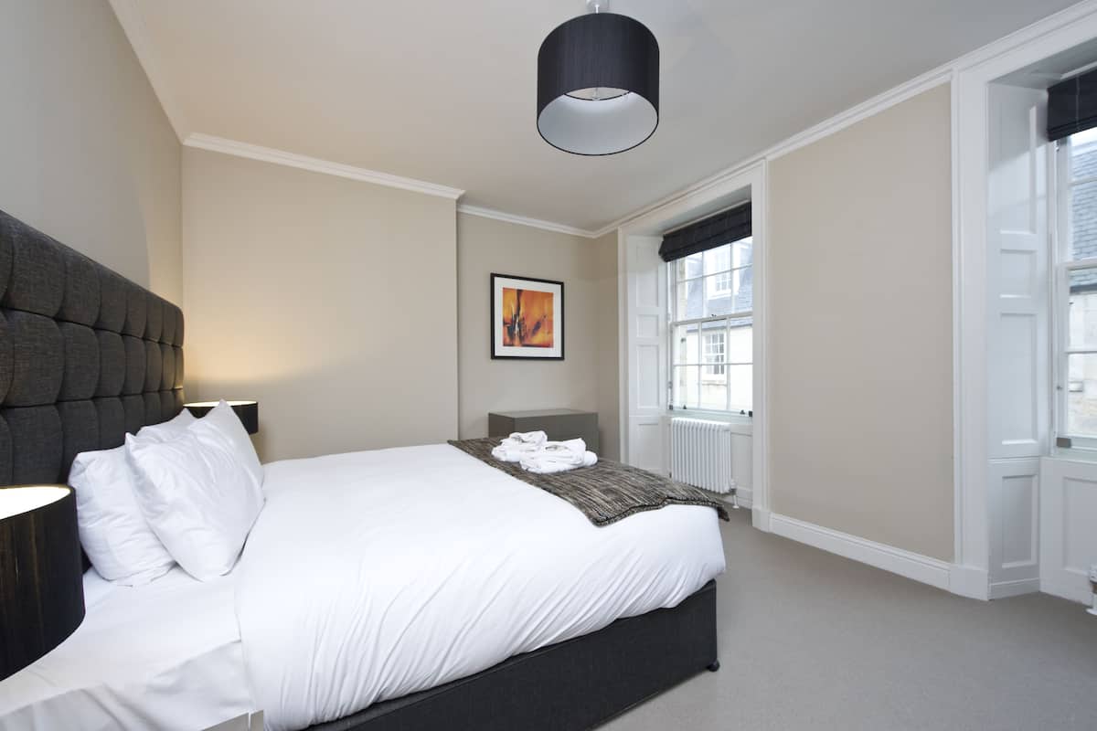 Une chambre à coucher avec un grand lit, une tête de lit, une table de chevet, une lampe, une fenêtre avec des stores et un tableau encadré au mur.