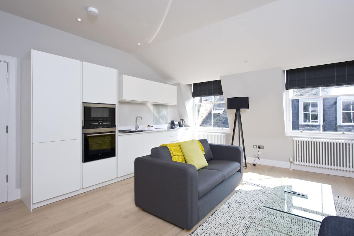 Une cuisine moderne avec des meubles blancs et un four encastré, un canapé gris agrémenté d’un coussin jaune et un lampadaire.