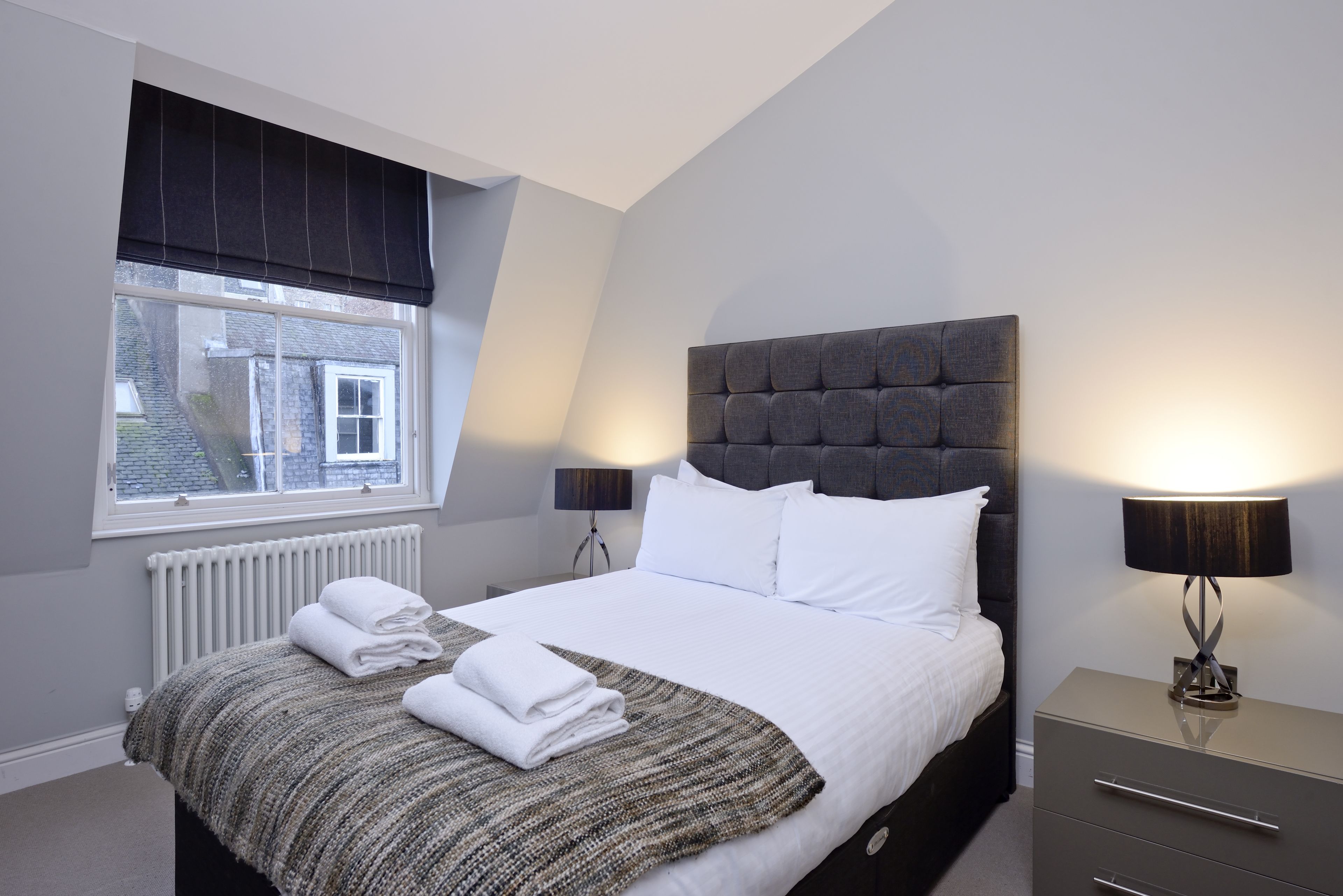 A neatly made bed with white linens, a grey headboard, and a bedside lamp.