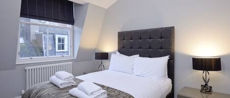 A neatly made bed with white linens, a grey headboard, and a bedside lamp.
