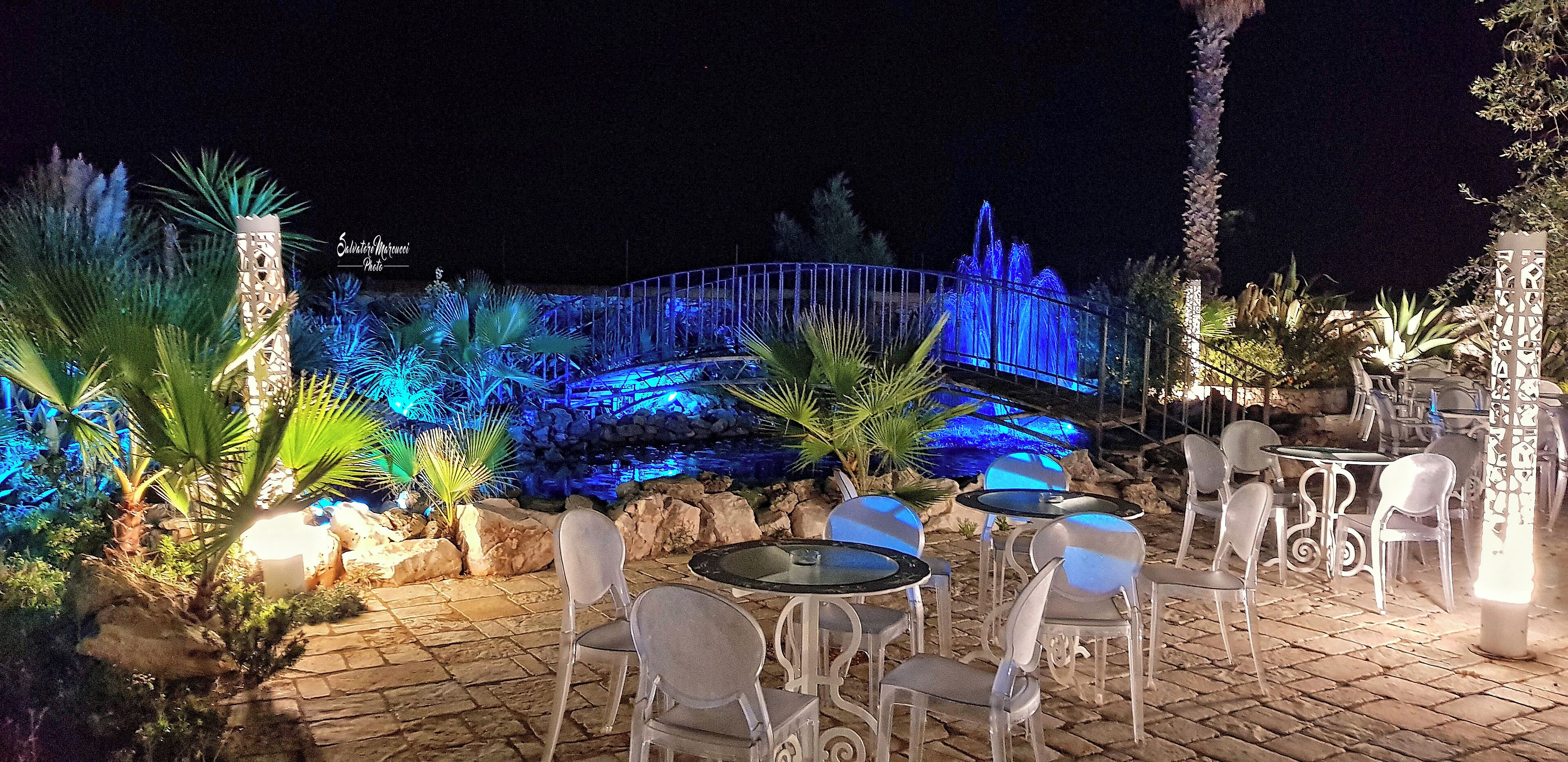 An outdoor patio area with tables and chairs, a decorative fence, and a water feature illuminated by blue lights.