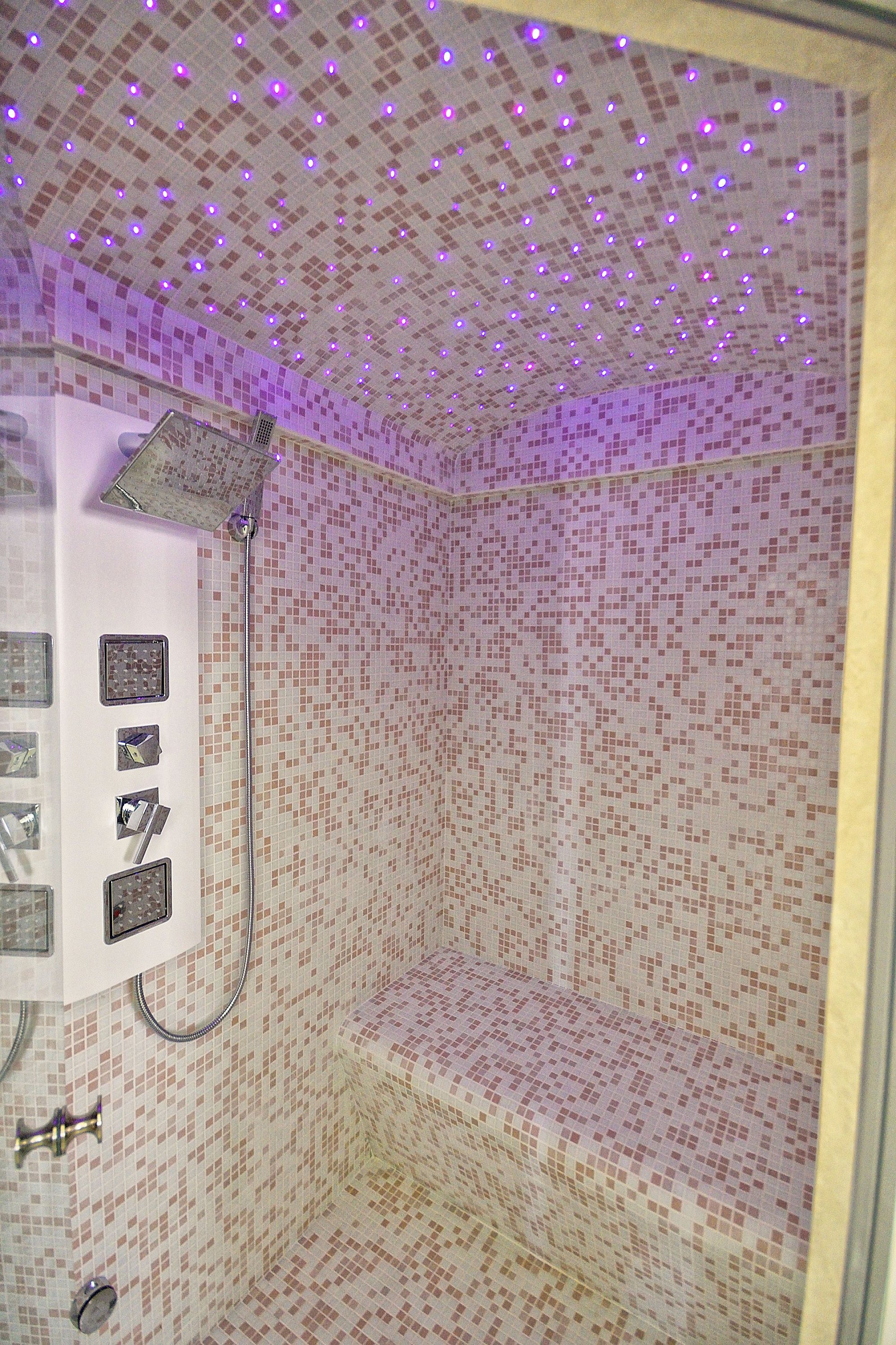Une douche avec un mur recouvert d’une mosaïque et un plafond éclairé de lumières violettes.