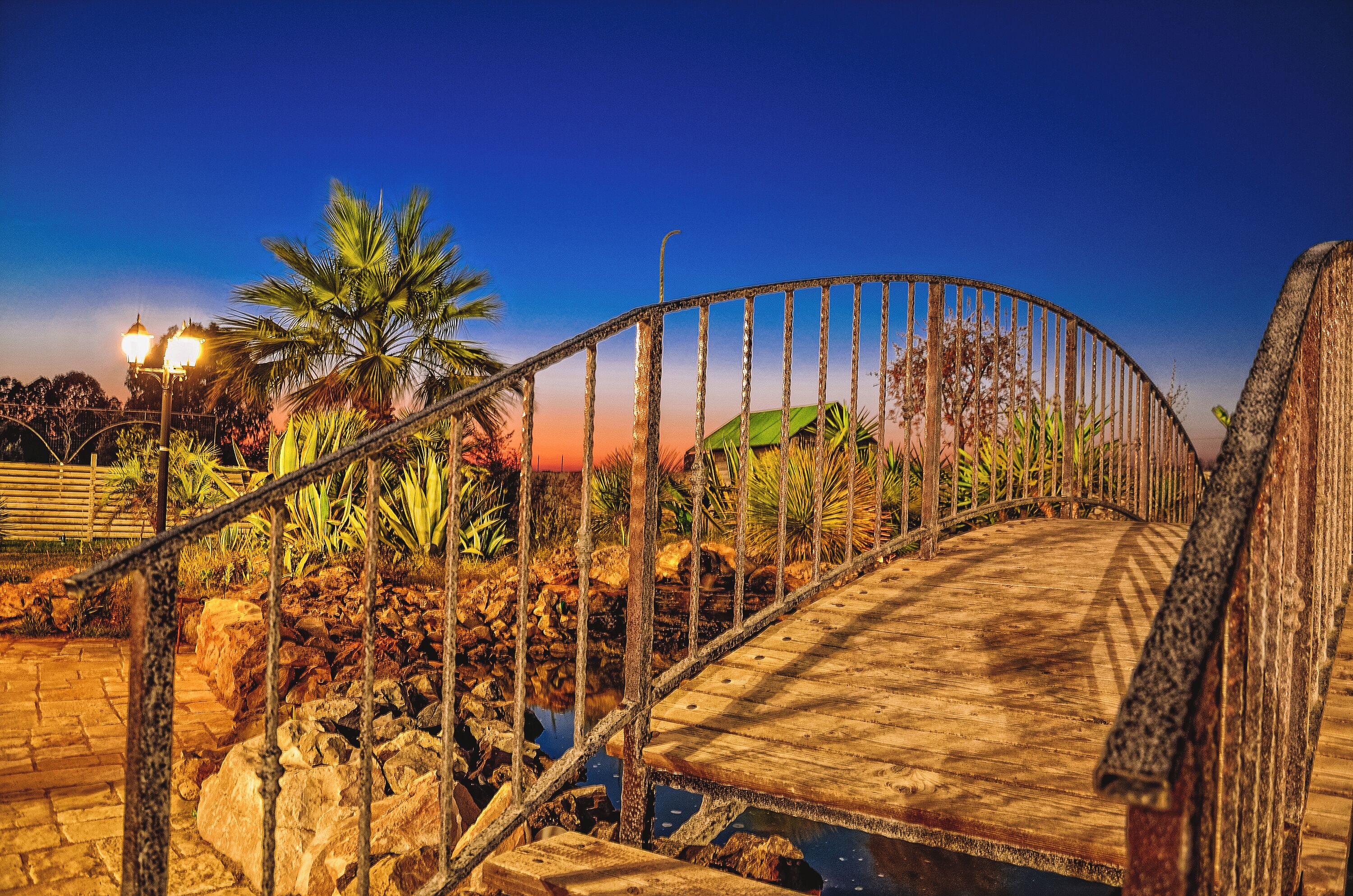 A wooden bridge with metal railings, surrounded by rocks and plants, under a clear sky.