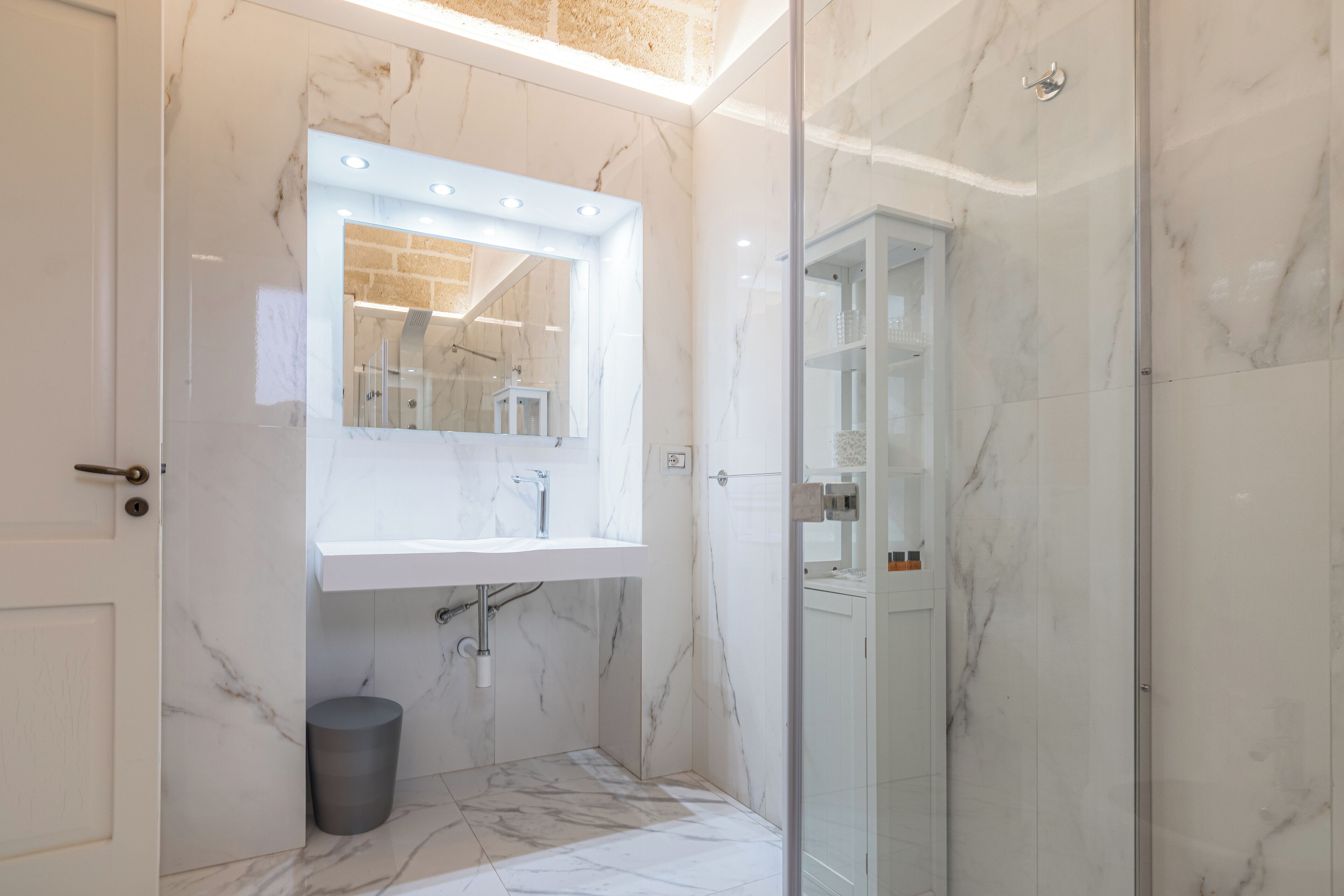 A modern bathroom with a marble sink, a glass shower enclosure, and a visible trash bin.