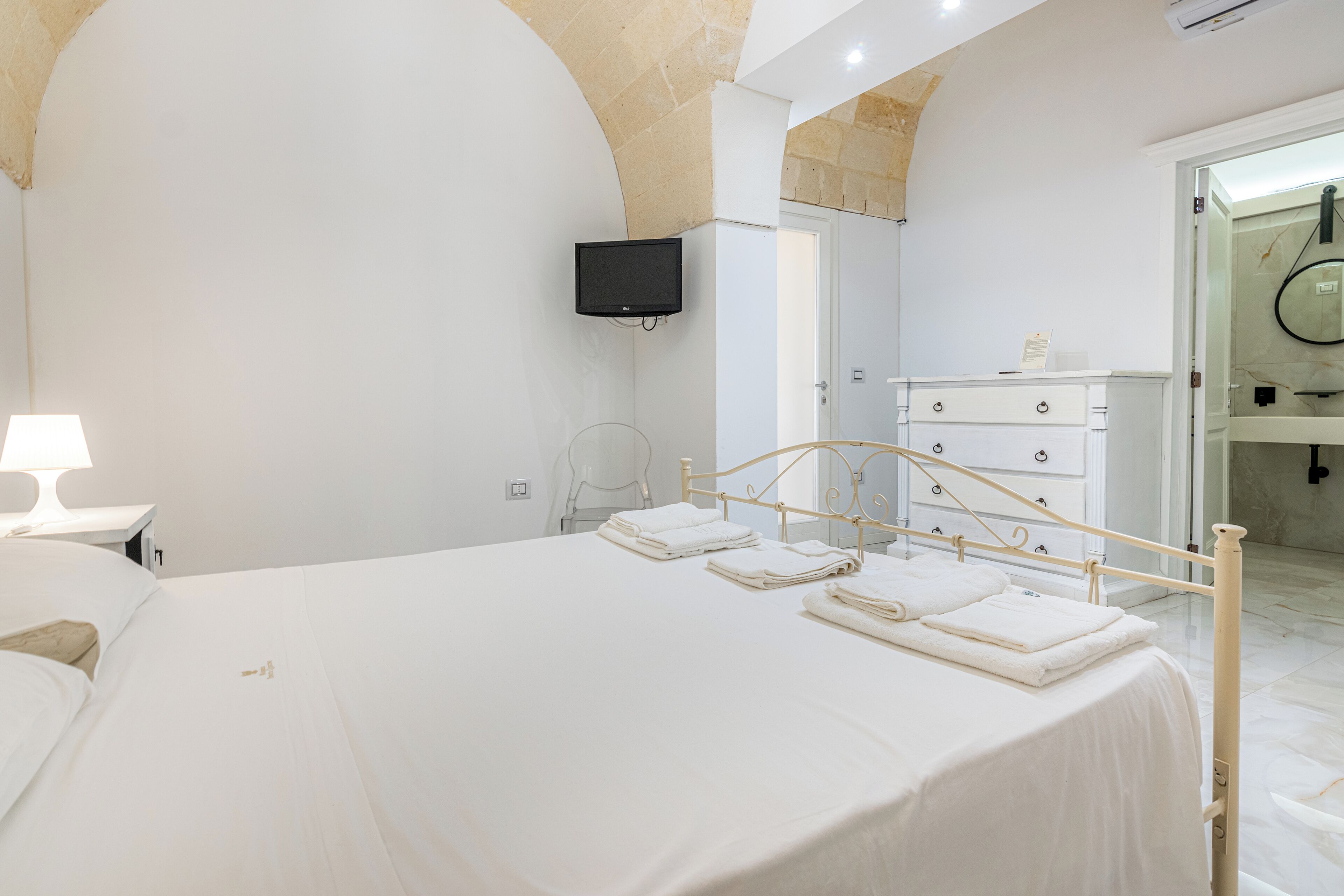 A bedroom with a bed, a television mounted on the wall, a dresser, and a bathroom visible through an open door.