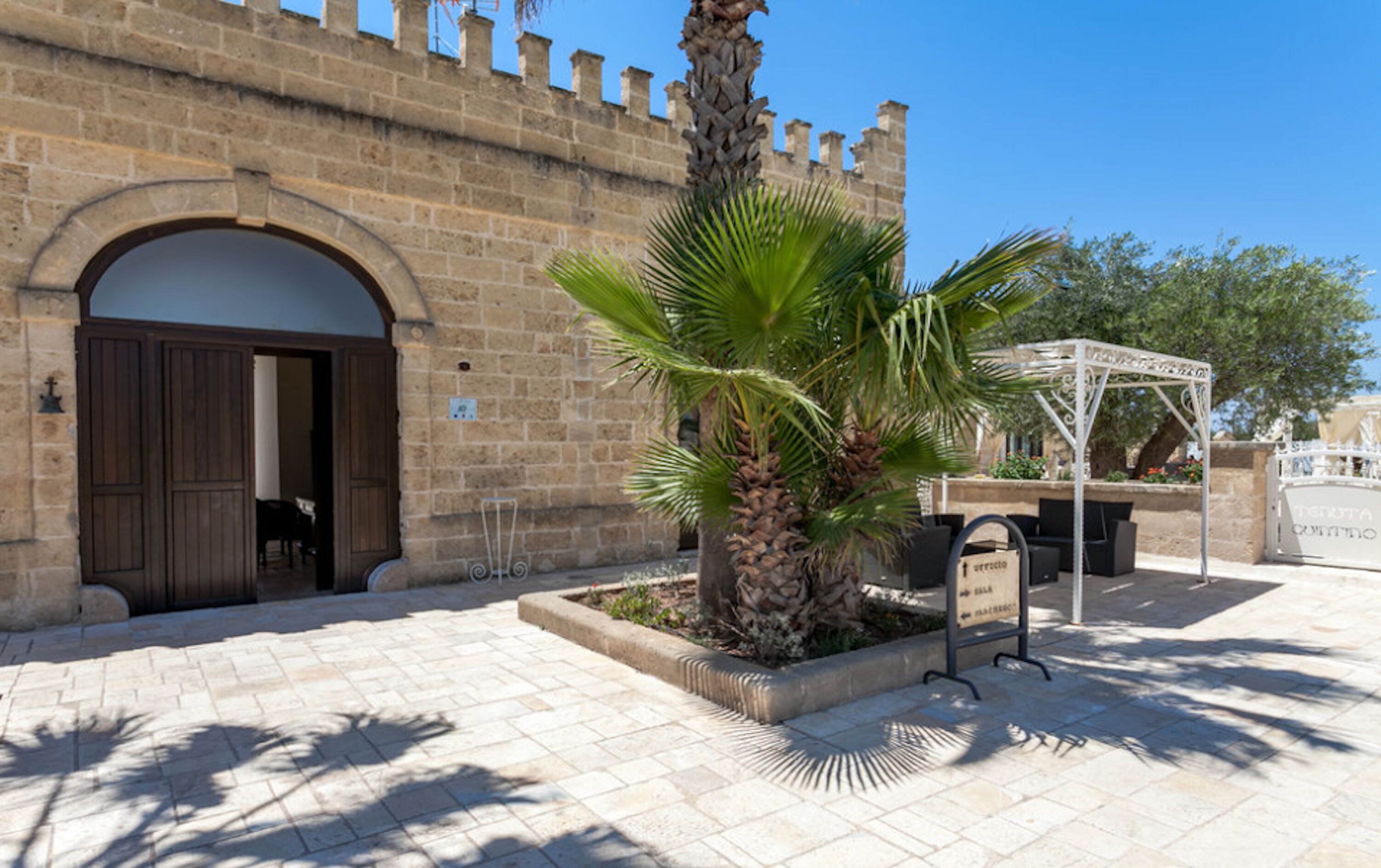 A courtyard with a palm tree, a stone building with an arched entrance, and outdoor seating.