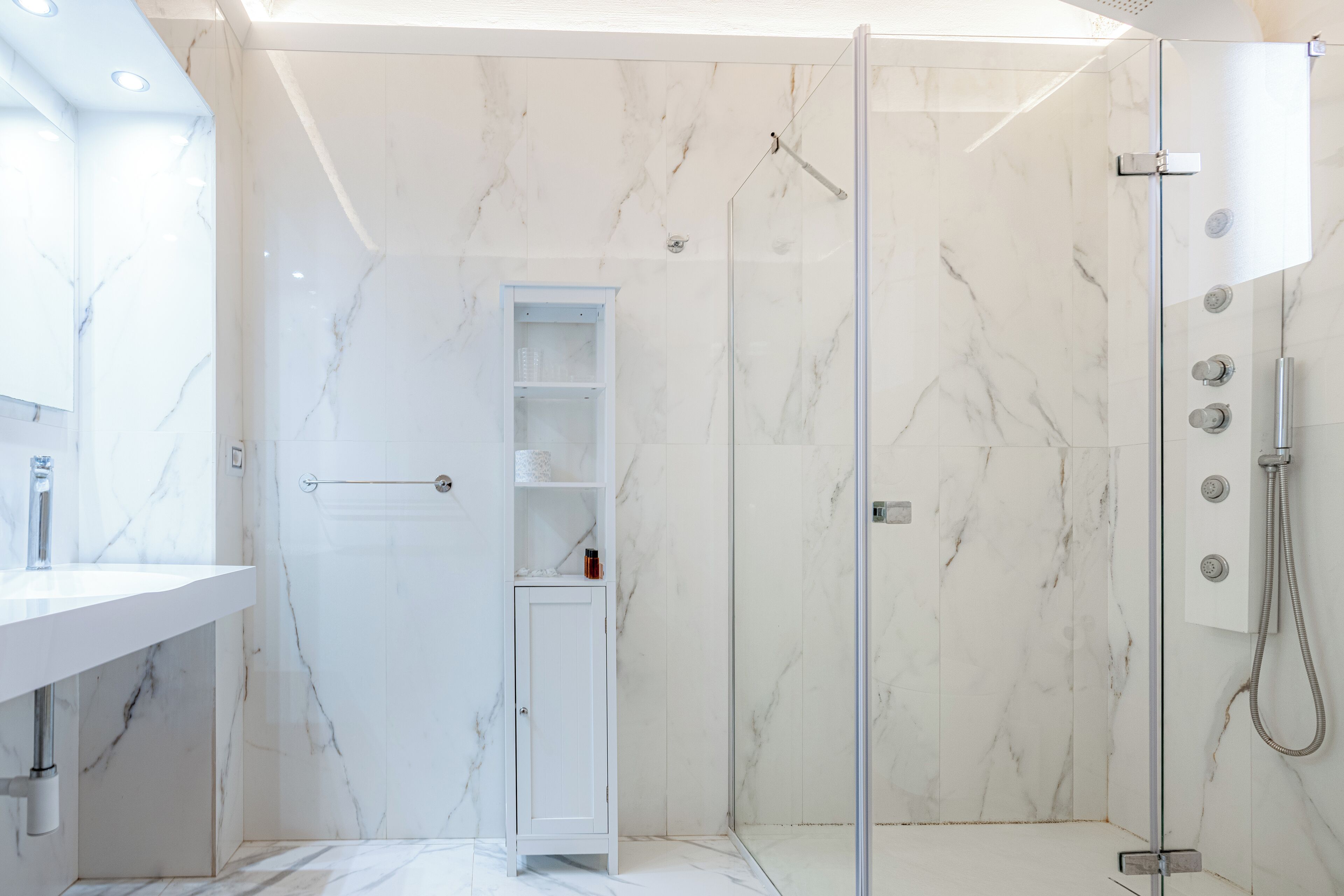 Un baño moderno con piso de mármol, ducha con mampara de vidrio, tocador blanco con lavamanos y un estante empotrado.
