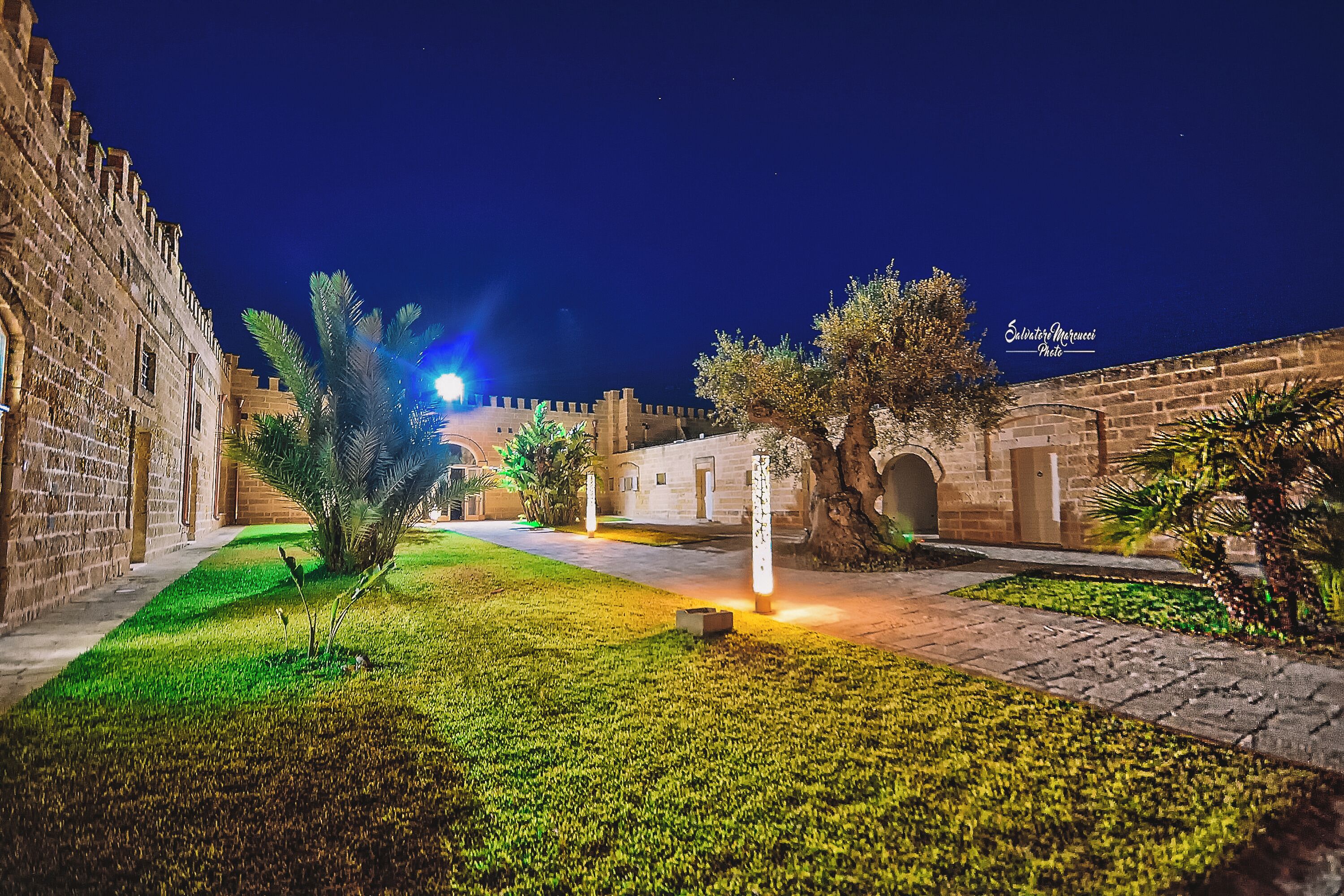 A courtyard with stone walls, palm trees, and a well-lit pathway.
