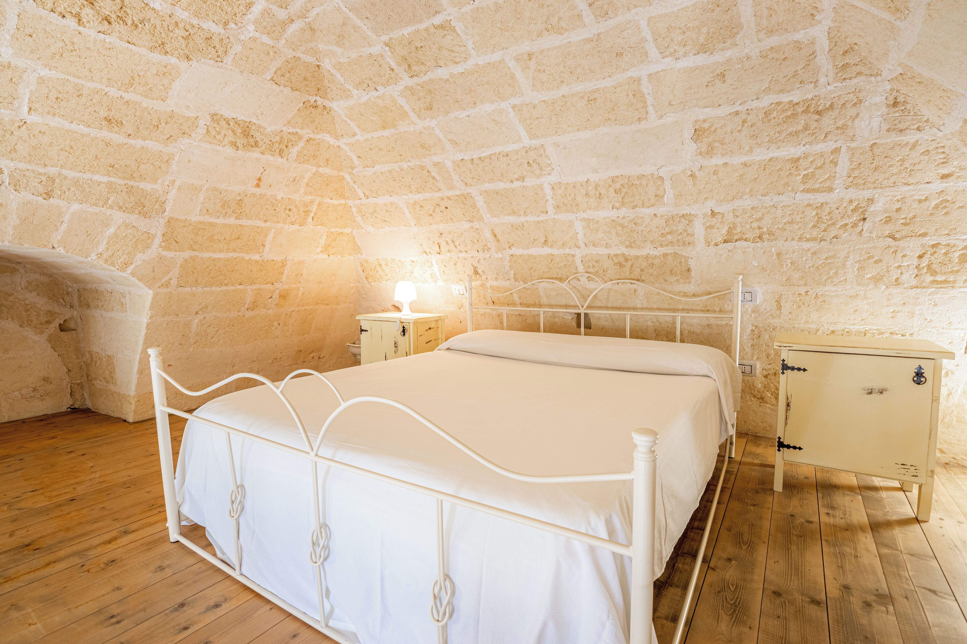 A bedroom with a white metal bed, wooden flooring, and a stone wall.