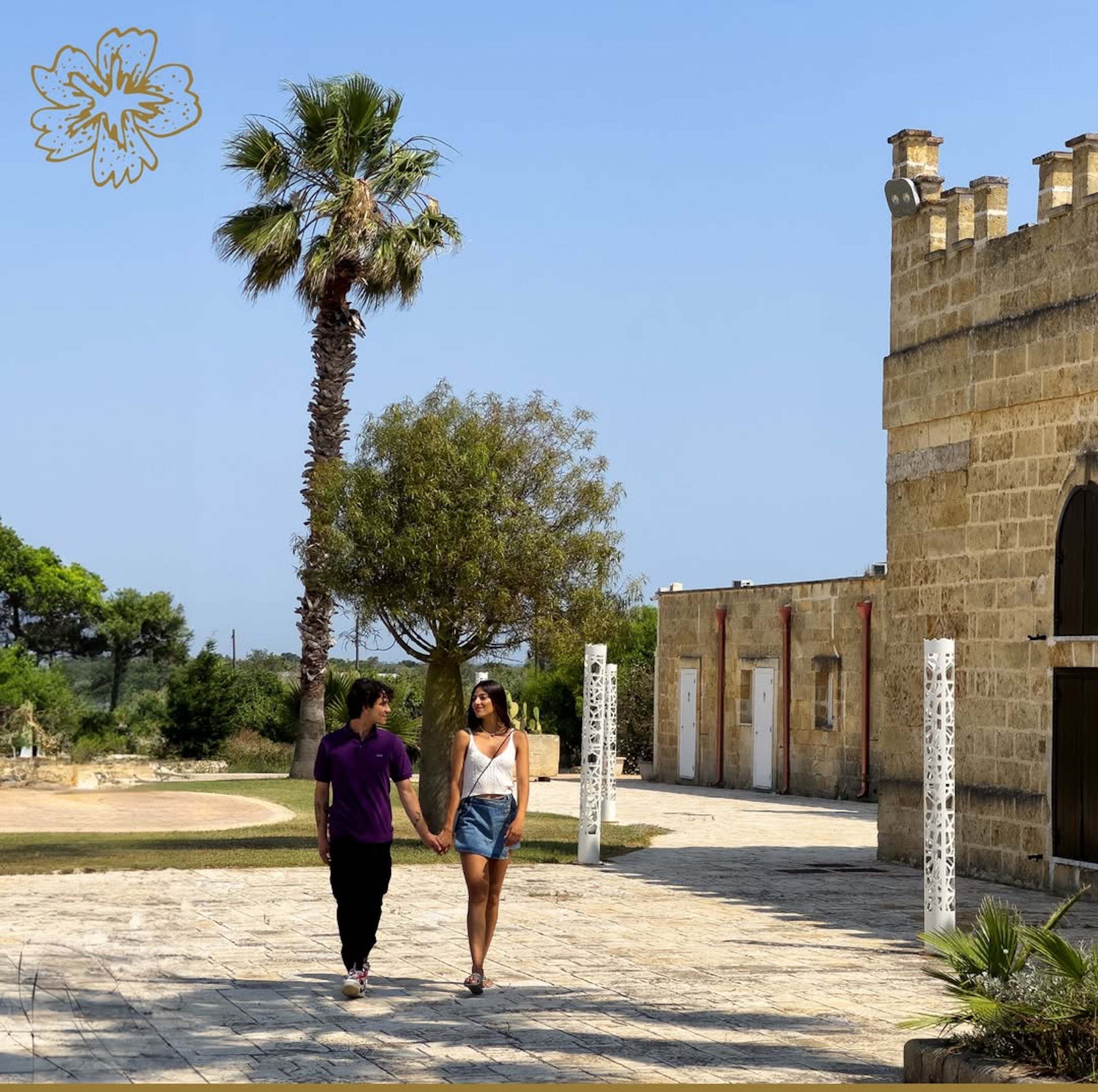 A couple walking on a paved path with a palm tree and a stone building in the background.