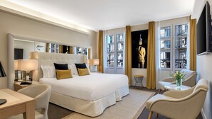 Habitación de hotel moderna con una cama grande, un escritorio, dos sillas, un televisor y vistas a la ciudad a través de amplias ventanas.