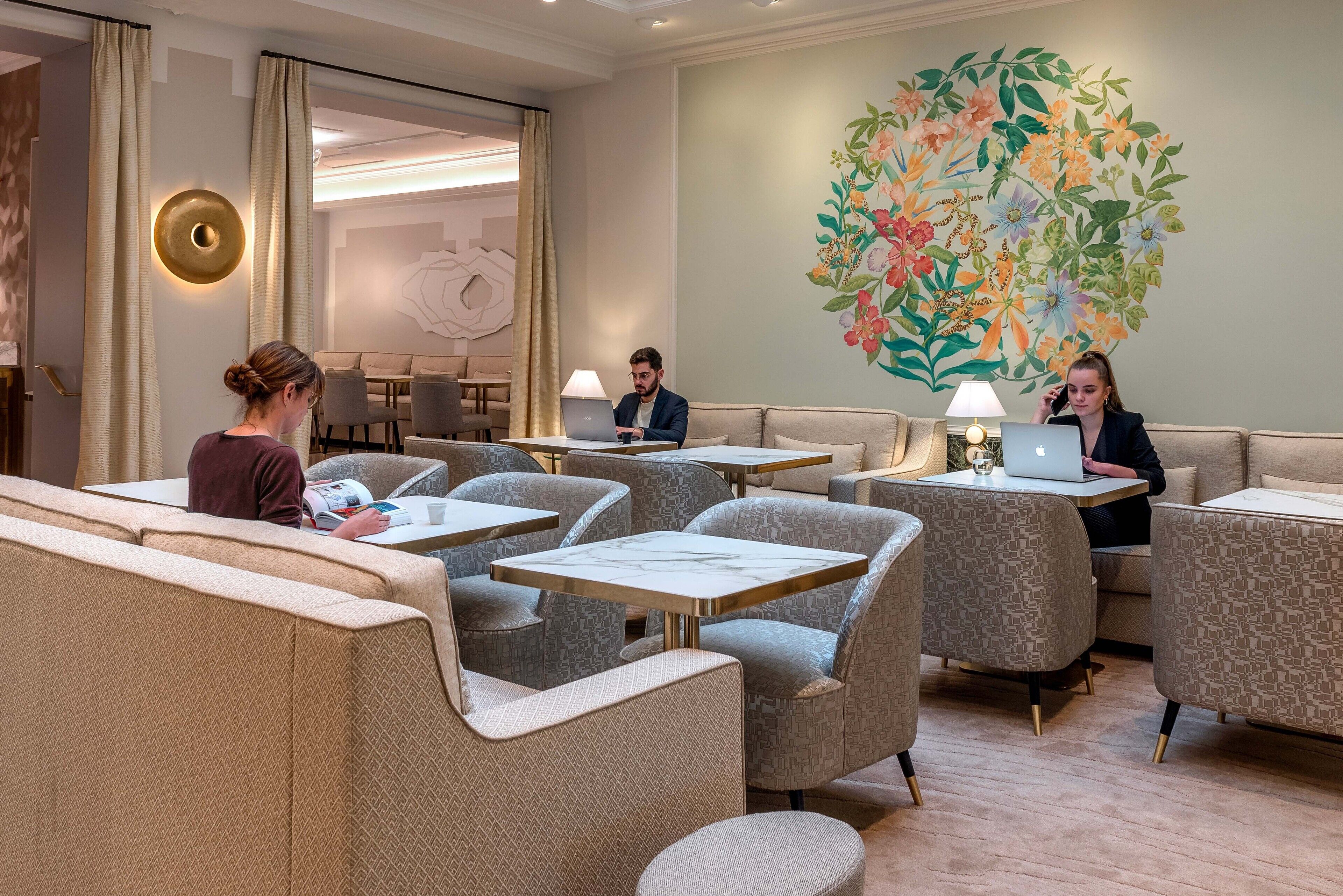 A modern lounge area with seating and tables, a large floral mural on the wall, and a round wall light.