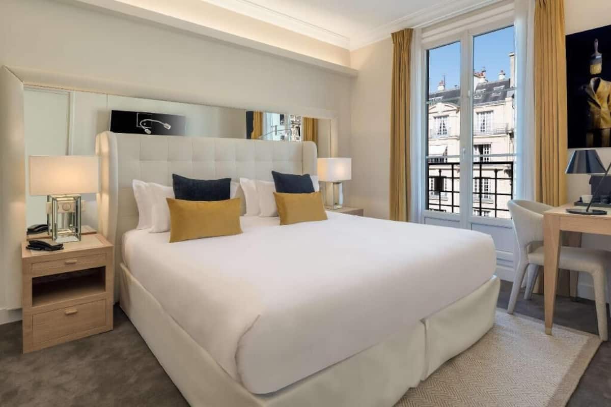 Habitación de hotel con una cama grande, mesitas de noche, un escritorio y vistas a edificios a través de una ventana.