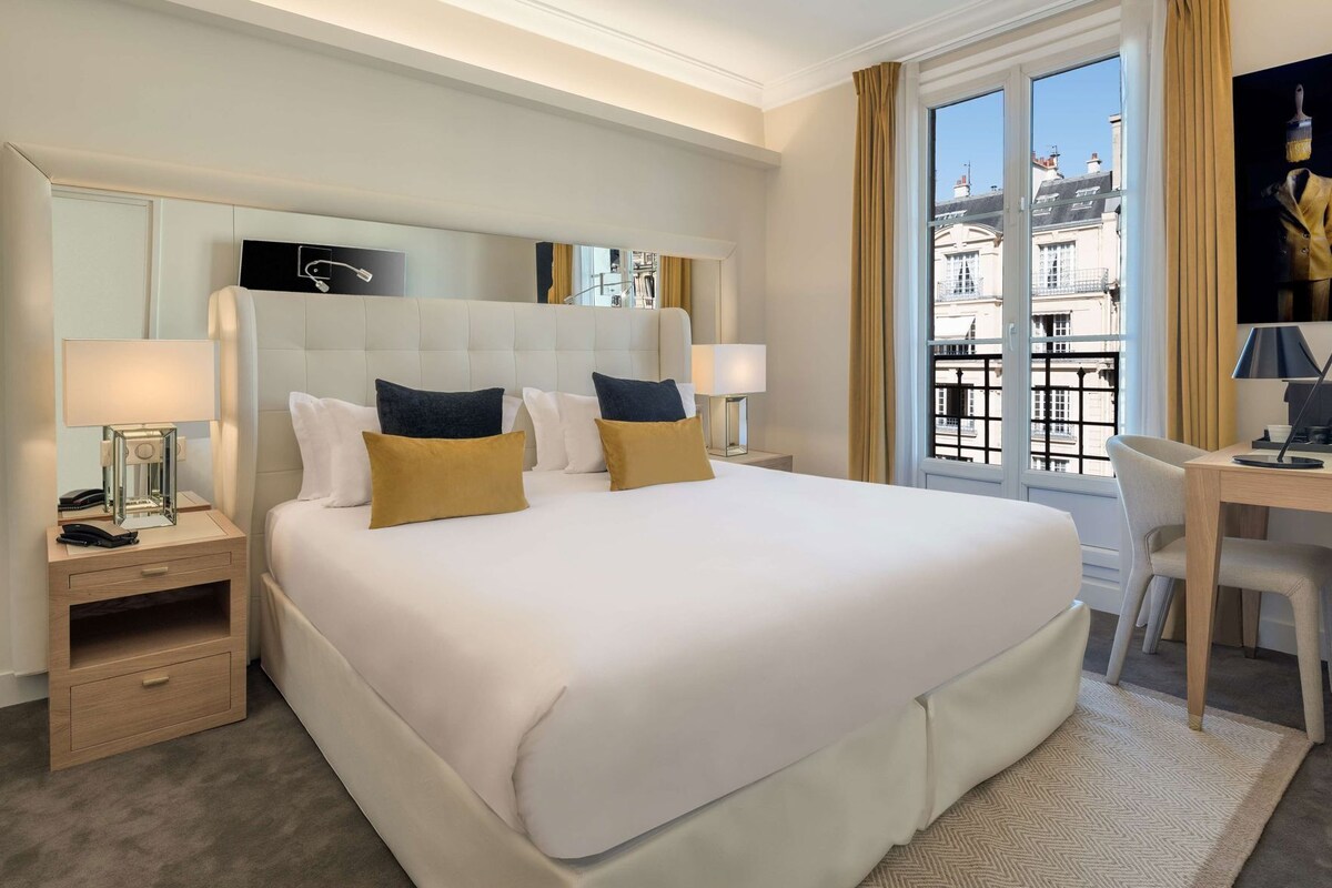 Une chambre d’hôtel avec un grand lit, des tables de chevet, un bureau et une vue sur les immeubles par la fenêtre.