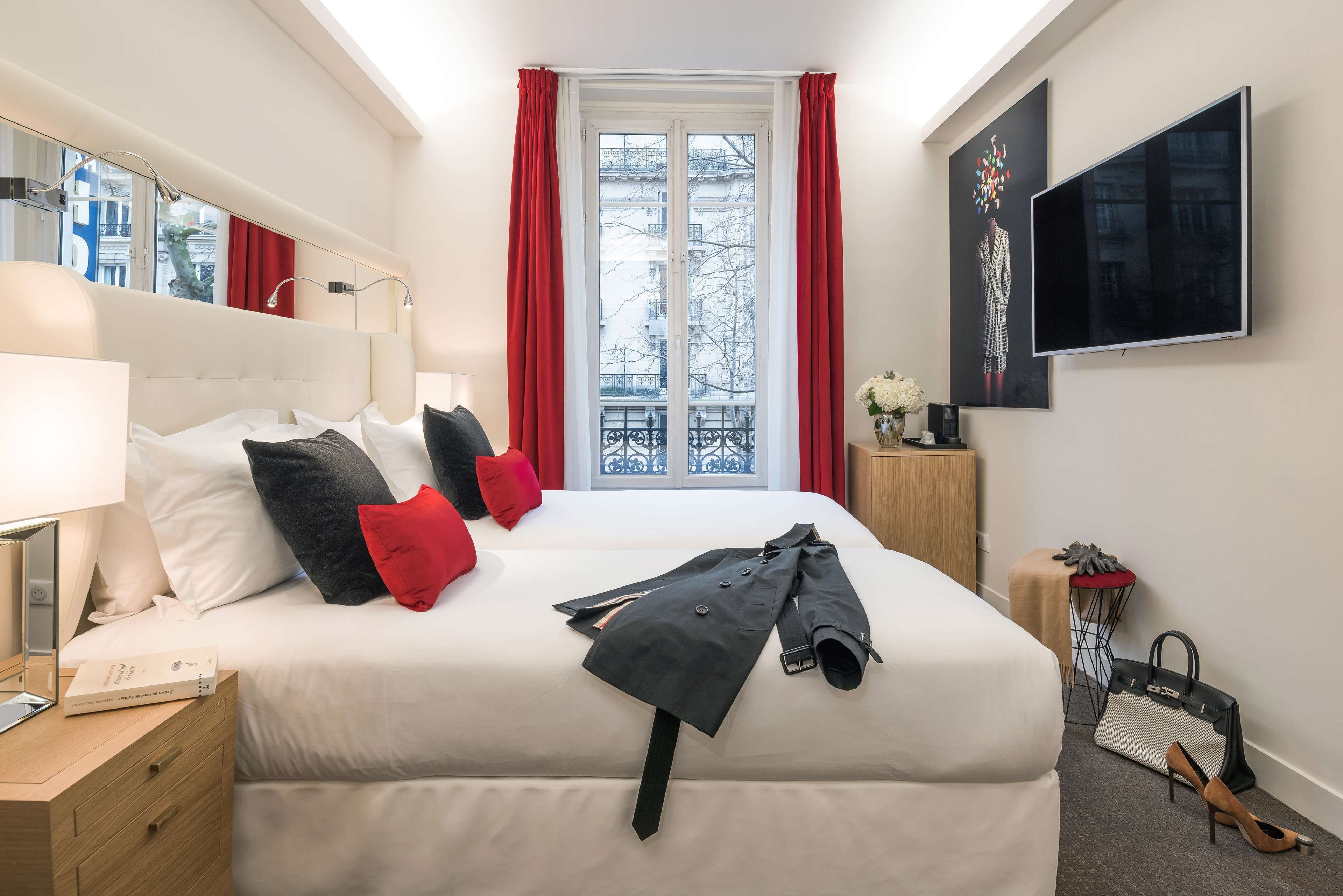 A hotel room with a bed, a bedside table with a lamp, a TV mounted on the wall, and a window with red curtains.