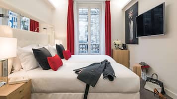 A hotel room with a bed, a bedside table with a lamp, a TV mounted on the wall, and a window with red curtains.