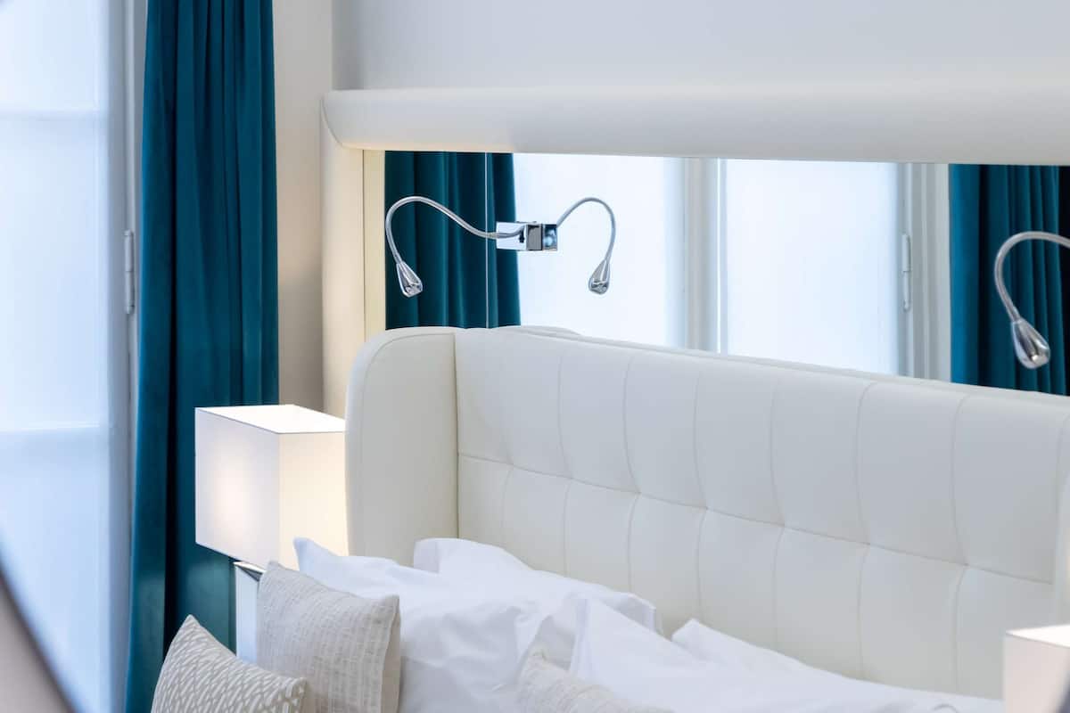Una habitación de hotel moderna con una cama blanca, varios almohadones, una mesita de noche y una lámpara.