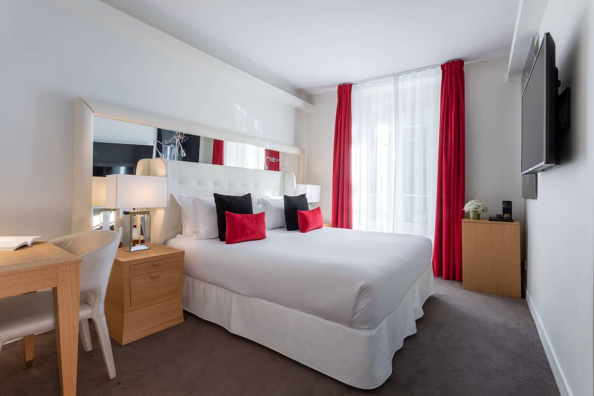 Habitación de hotel con una cama grande, un escritorio, una silla, una mesita de noche, un espejo, un televisor y cortinas rojas.