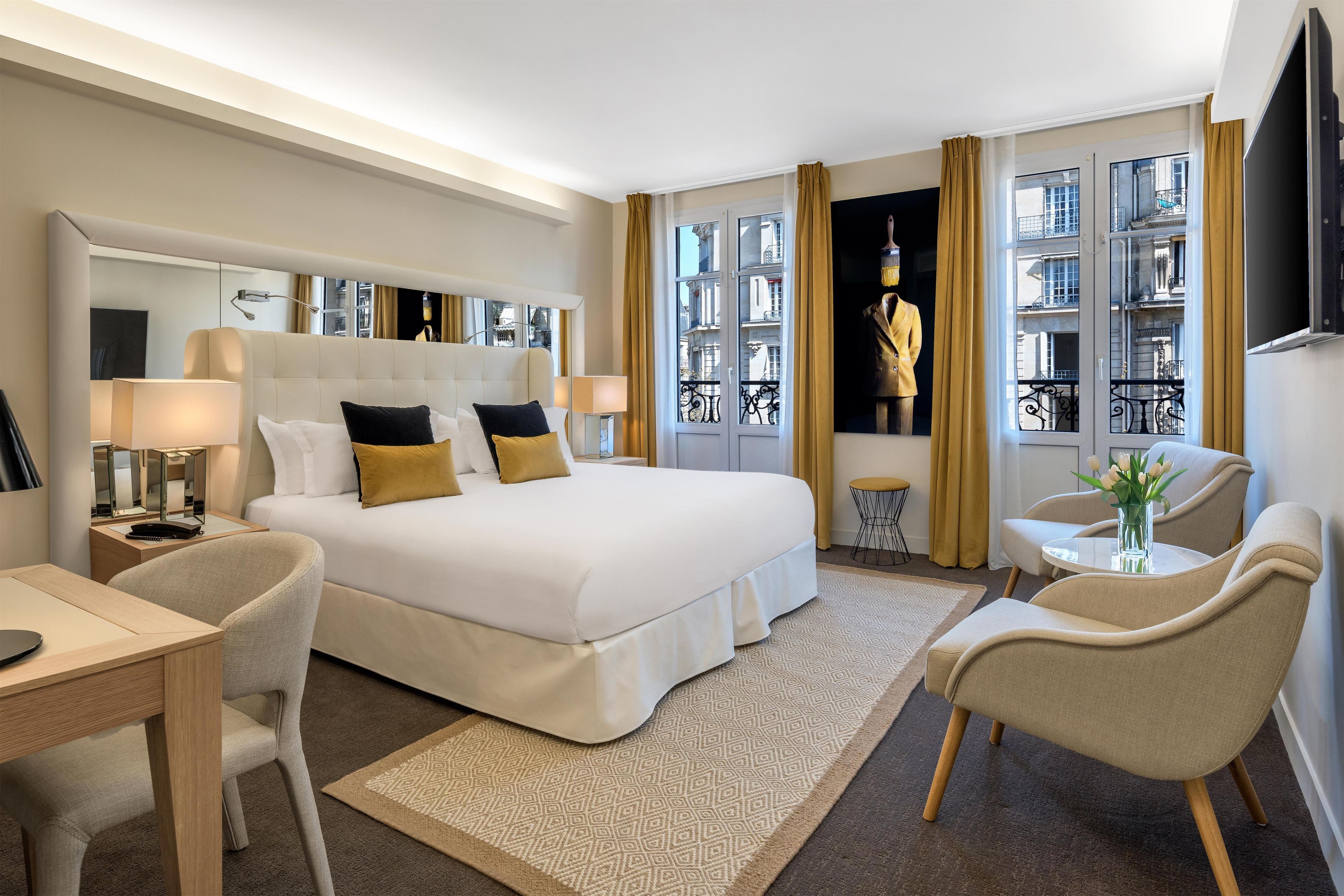 Ein Hotelzimmer mit einem großen Bett, einem Schreibtisch, einem Stuhl, einem kleinen Tisch mit einer Vase und einem Fenster mit Vorhängen.