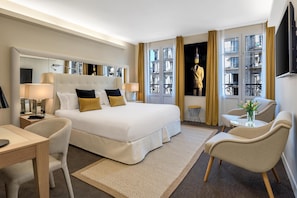 Een hotelkamer met een groot bed, een bureau, een stoel, een klein tafeltje met een vaas en een raam met gordijnen.