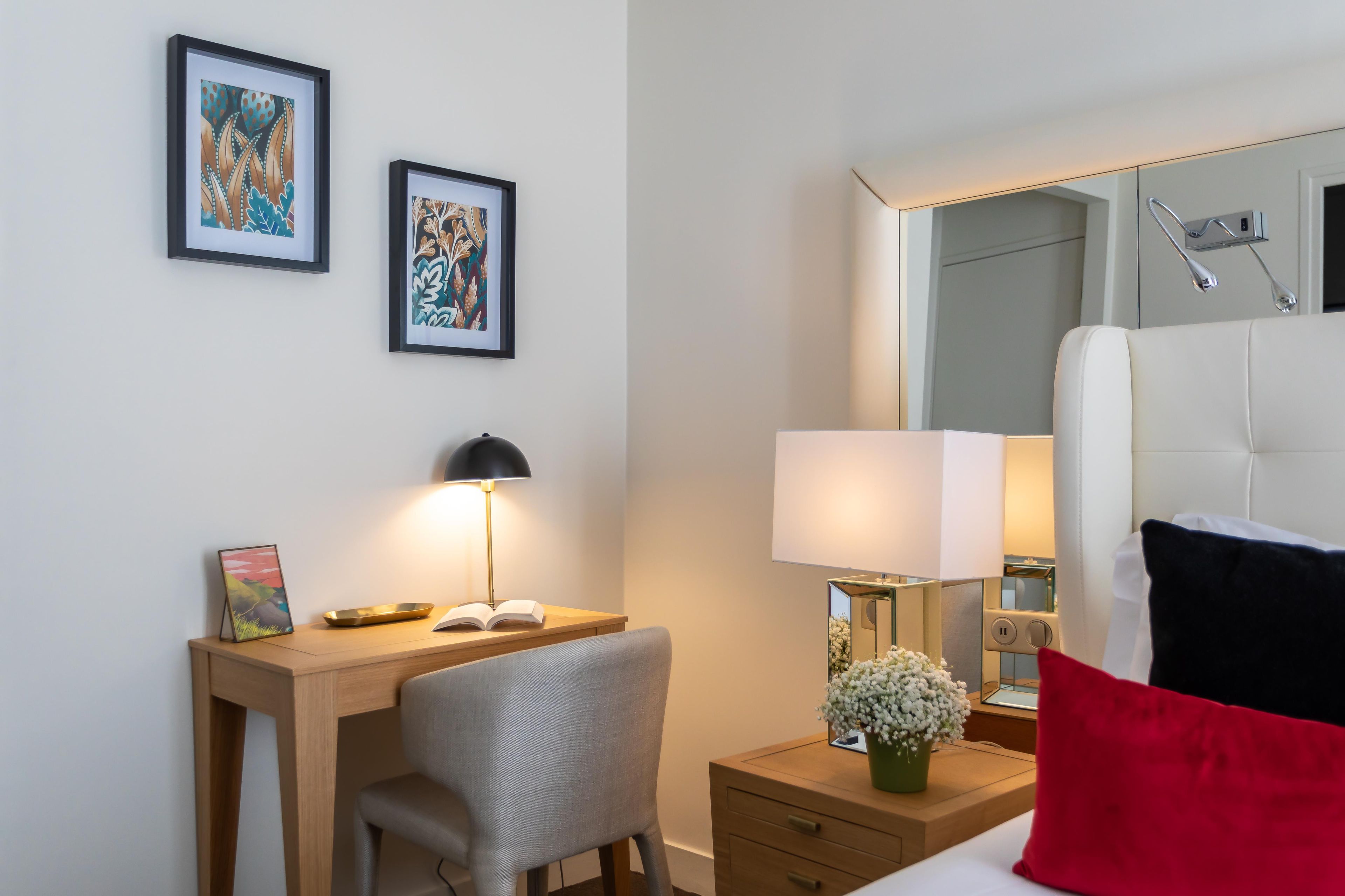 A hotel room with a desk, chair, bedside table with a lamp, a mirror, and a bed with pillows.