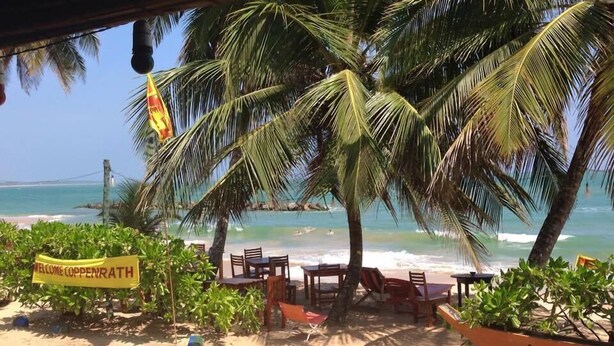 A beachside seating area with a 'Welcome to Copperrath' sign, palm trees, and a view of the ocean. - Coppenrath (Tangalle)