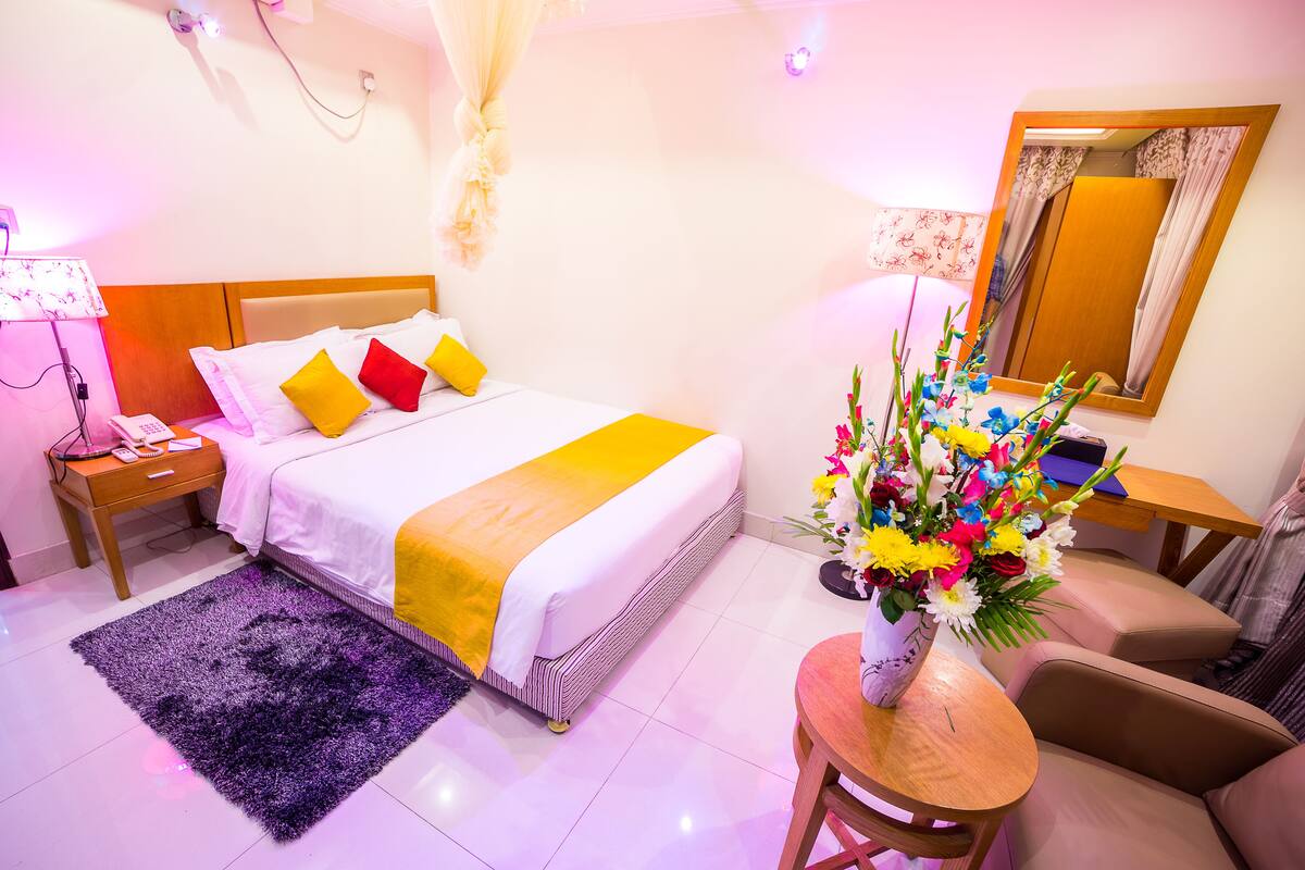 A hotel room with a bed, bedside table, lamp, chair, and a vase of flowers.