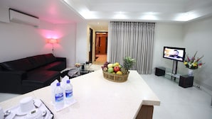 A modern living room with a black leather sofa, a glass coffee table, a fruit basket, a TV, and a small table with a tray of white cups and a bottle of water. - Nascent Gardenia Suites (Dhaka)