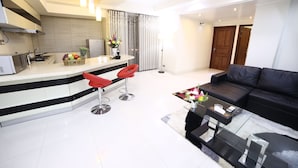 A modern kitchen with a white countertop, red bar stools, and a dining area with a black leather sofa and glass coffee table. - Nascent Gardenia Suites (Dhaka)