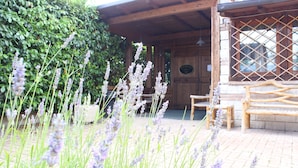 Eine Holzhütte mit einer Ziegelterrasse, umgeben von üppiger Vegetation und Lavendelstauden.
