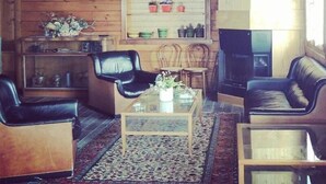 Ein gemütliches Wohnzimmer mit Holzwänden, einem Fernseher, der über einem Regal mit Dekotellern angebracht ist, und einem Kamin. Zwei Ledersofas, ein Couchtisch und ein Teppich liegen auf dem Boden.