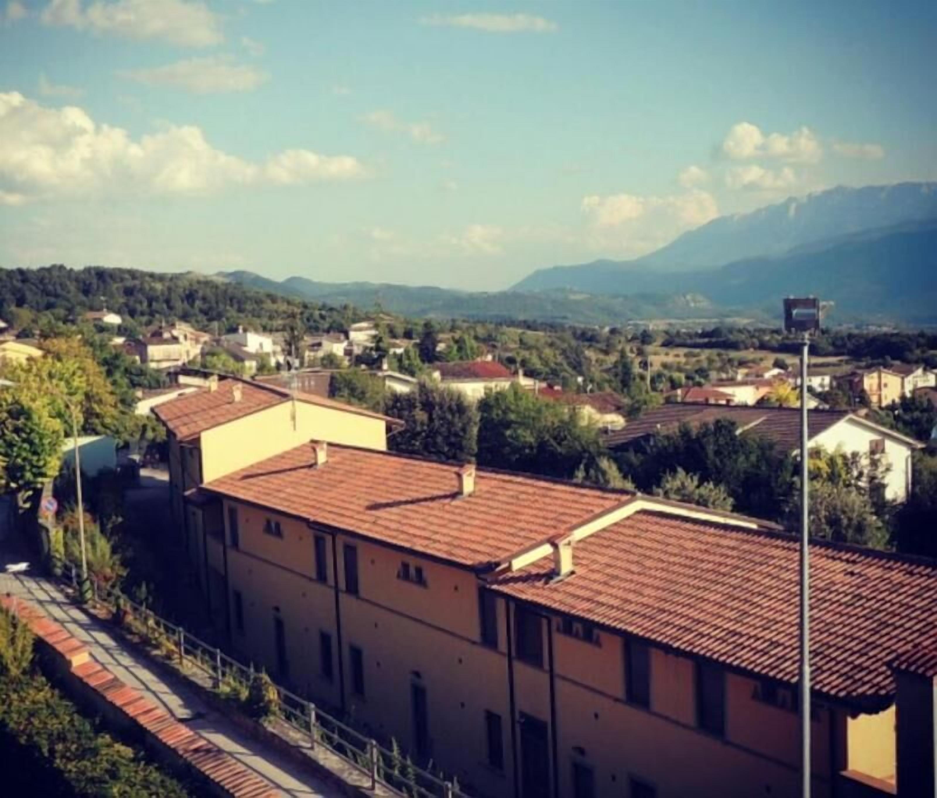 A residential area with terracotta-roofed buildings, a paved walkway, and a mountainous backdrop.