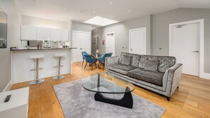 A modern living room with a grey sofa, a glass coffee table, and a dining area with blue chairs. - Destiny Scotland - St. Andrew Square Apartments (Edinburgh)