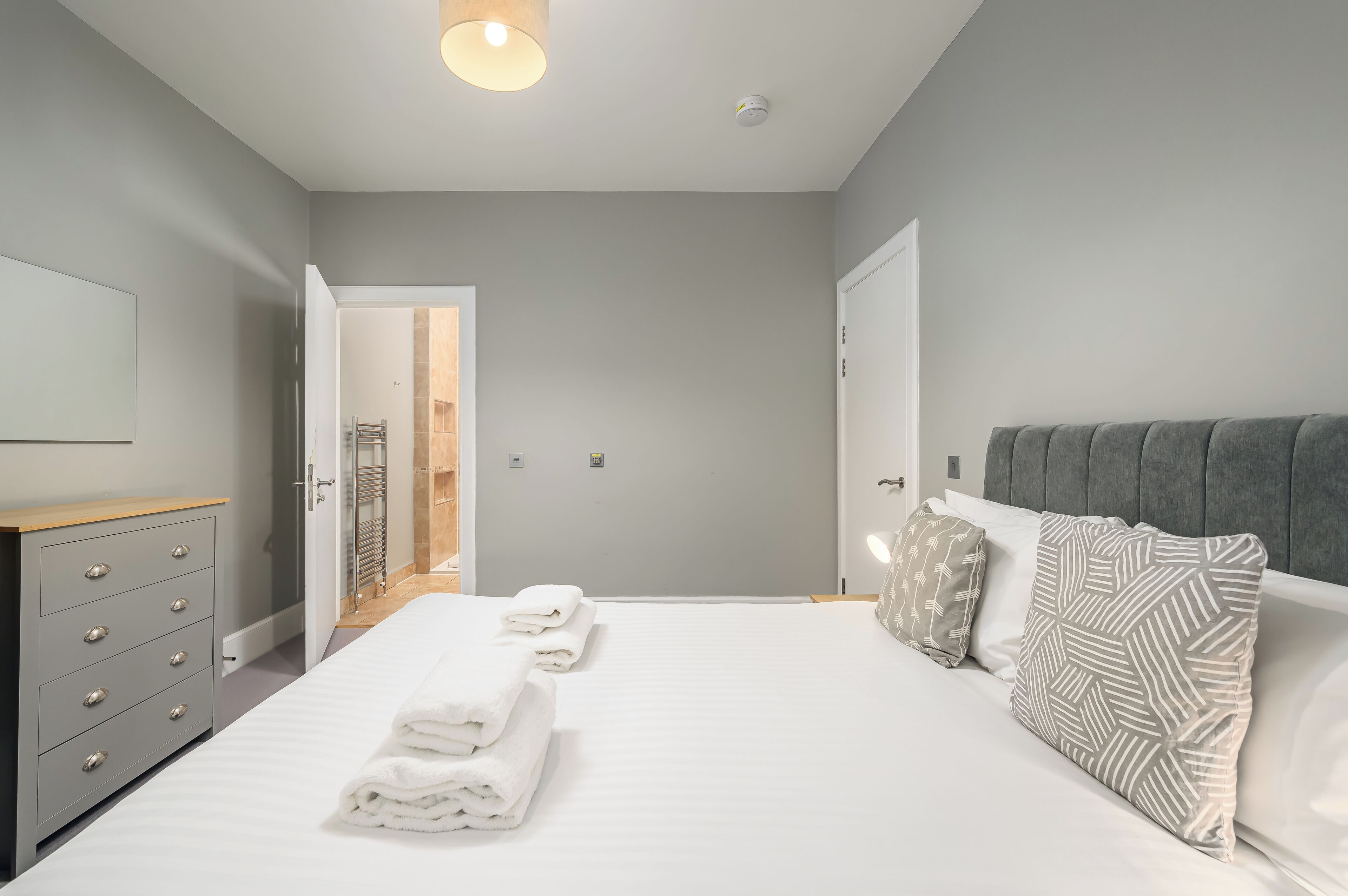 A modern bedroom with a large bed, grey walls, a wooden dresser, and a grey upholstered headboard.