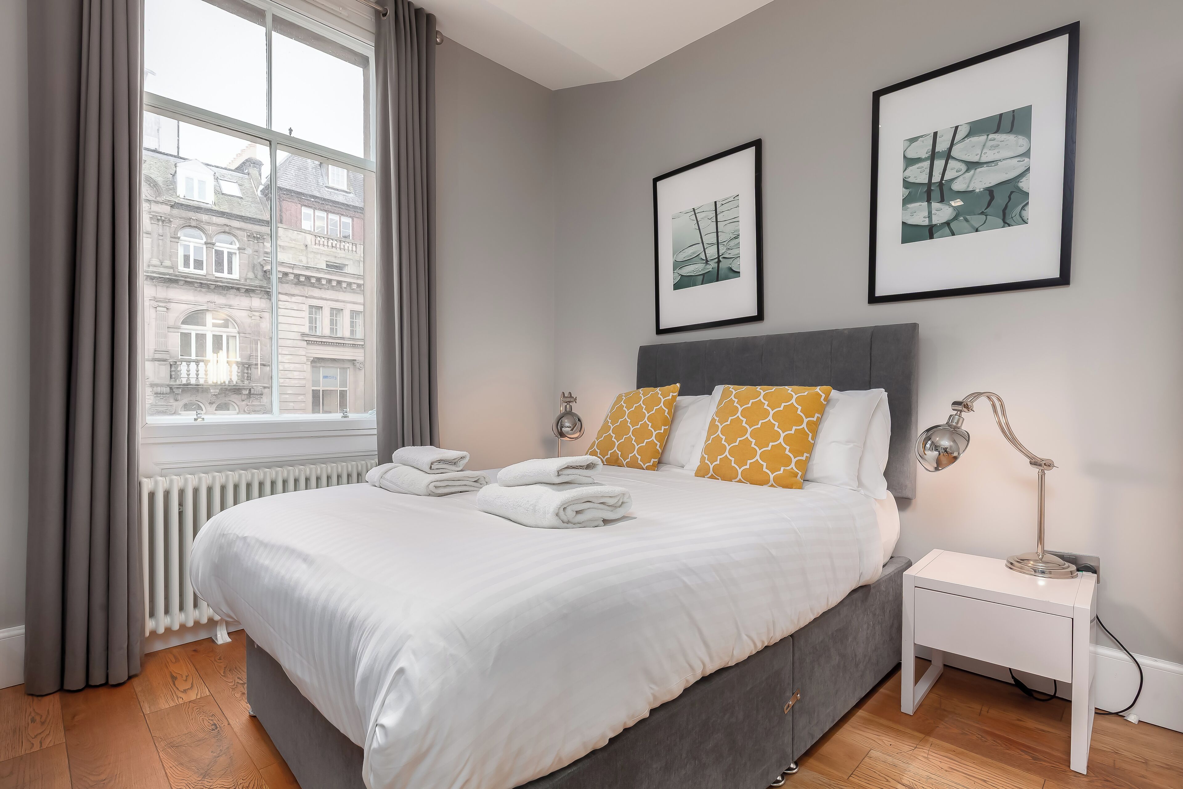 A neatly made bed with white linens and two pillows, a bedside table with a lamp, and two framed pictures on the wall.