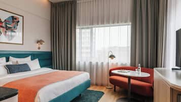 A hotel room with a bed, a desk, a chair, a table, a lamp, and a window with curtains.