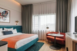 Une chambre d’hôtel avec un lit, un bureau, une chaise, une table, une lampe et une fenêtre avec des rideaux.