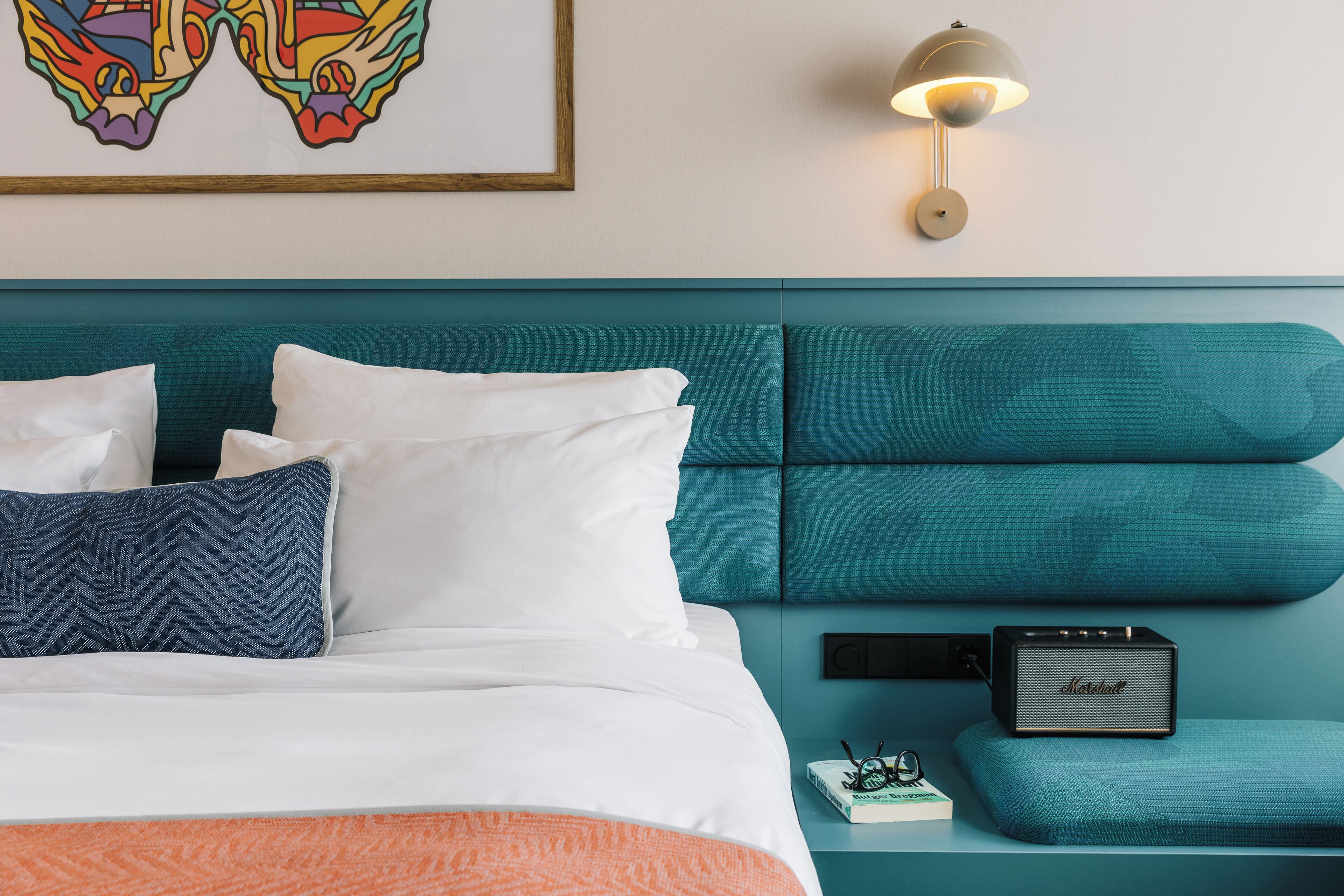 A neatly made bed with white and blue pillows, a teal headboard, a wall-mounted lamp, and a small speaker on a nightstand.