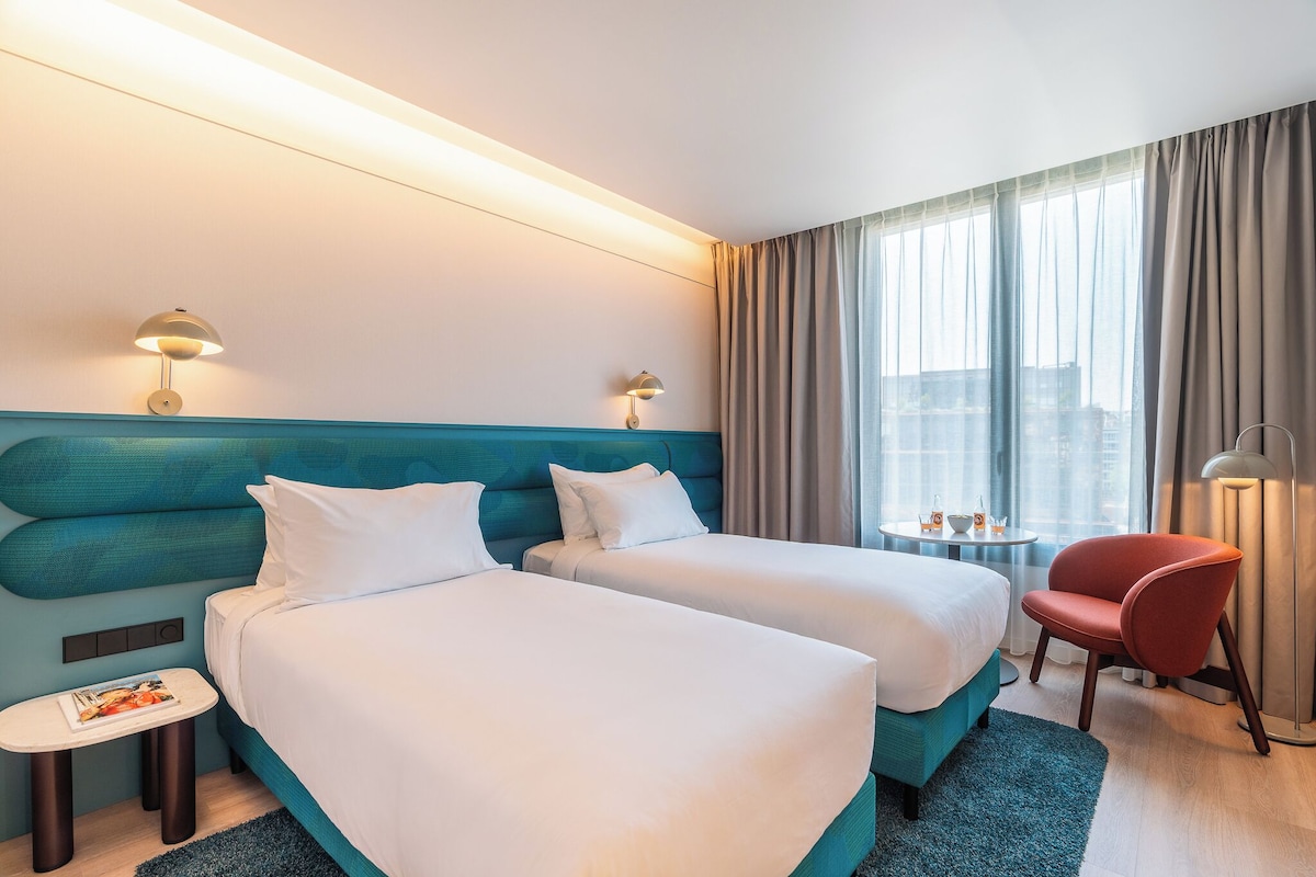 Een hotelkamer met twee bedden, een tafeltje, een stoel en uitzicht op de stad door grote ramen.