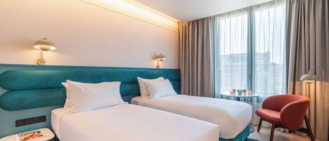 Habitación de hotel con dos camas, una mesita, una silla y vista de la ciudad a través de amplios ventanales.