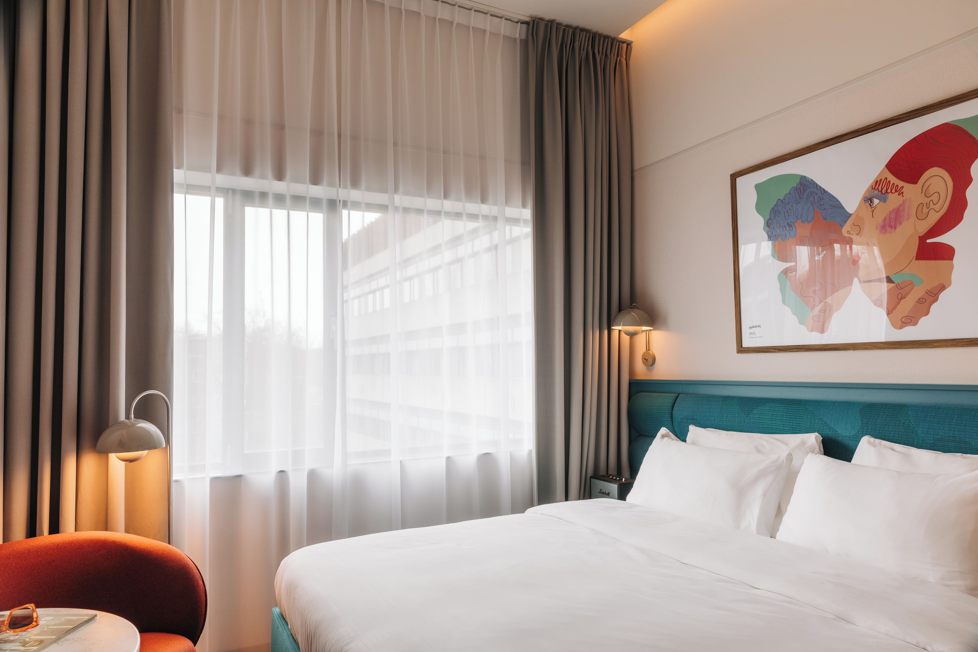 A hotel room with a large bed, a red chair, a nightstand with a lamp, and a window with sheer curtains.