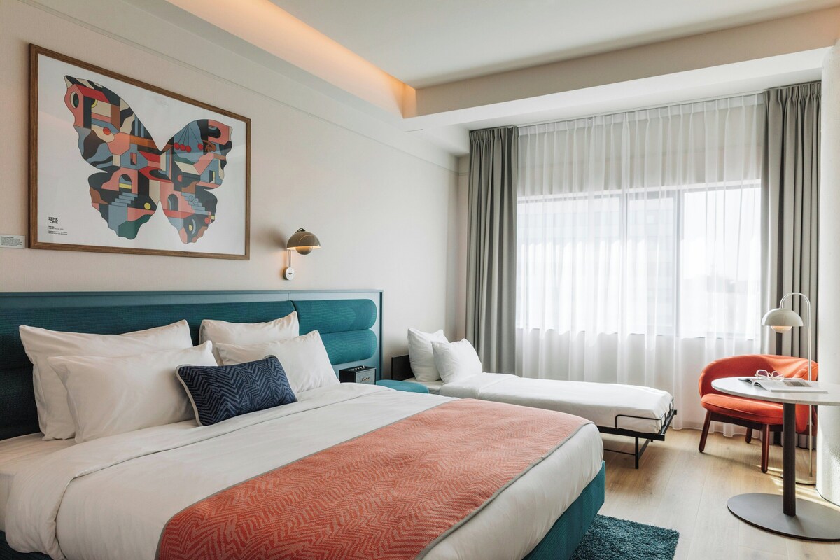 Een hotelkamer met een groot bed, een kleiner bed, een bureau met stoel, een raam met gordijnen en een wanddecoratie.
