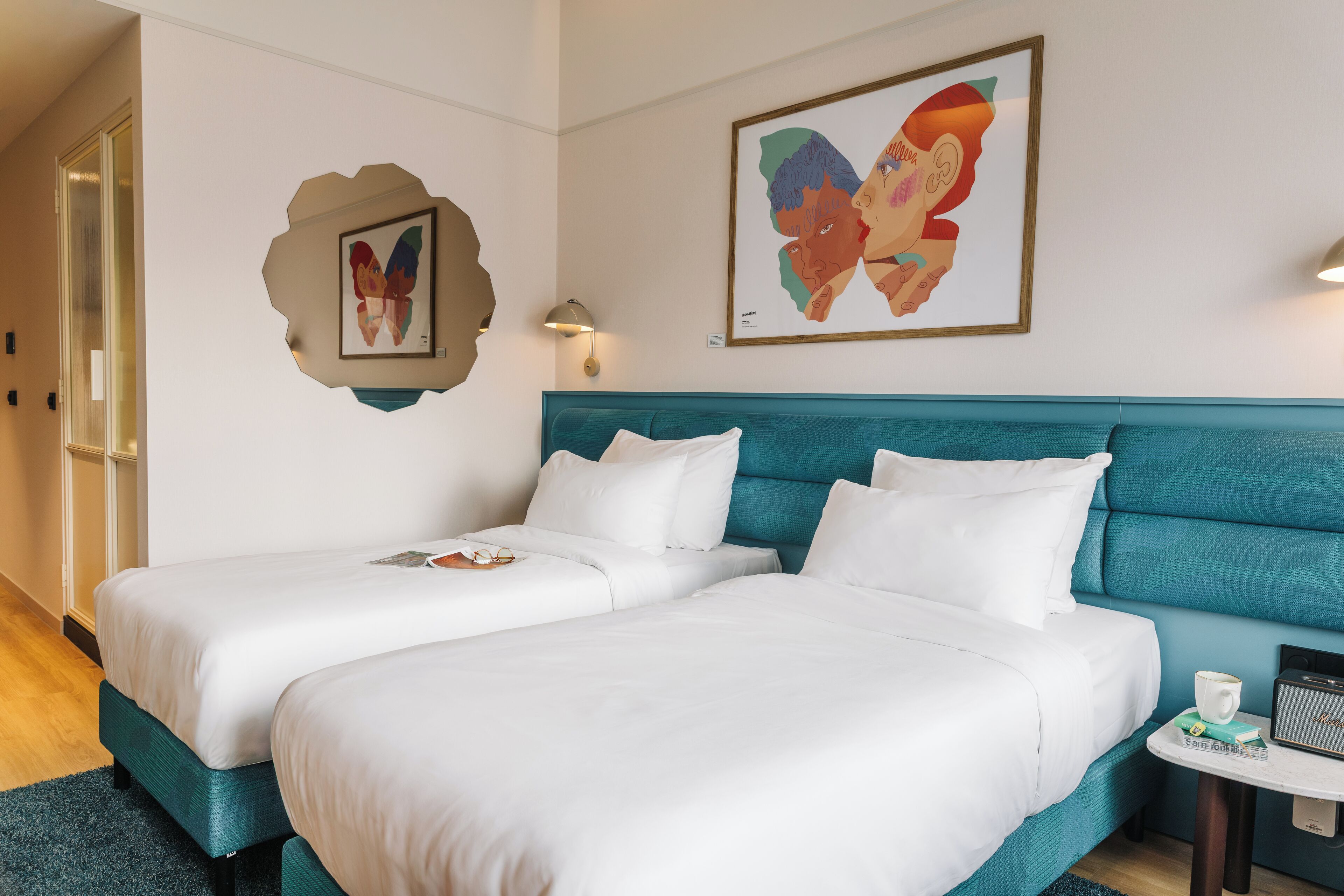 Une chambre d’hôtel avec deux lits, une tête de lit de couleur turquoise, une œuvre d’art représentant un papillon encadrée, une petite table avec une radio et une porte dotée de panneaux en verre dépoli.